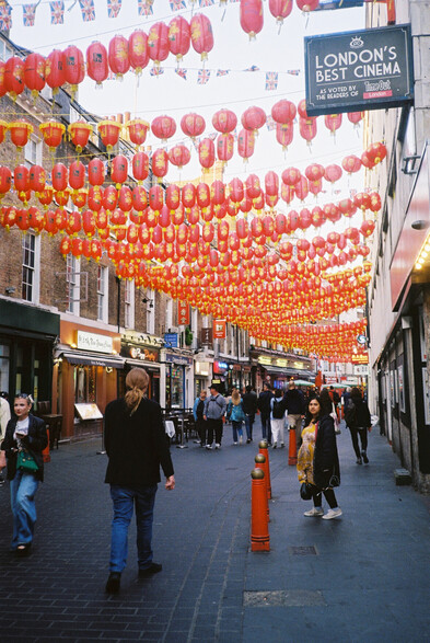 (color): Chinatown, London. A crowded street with many red lanterns hanging between the buildings above the street.