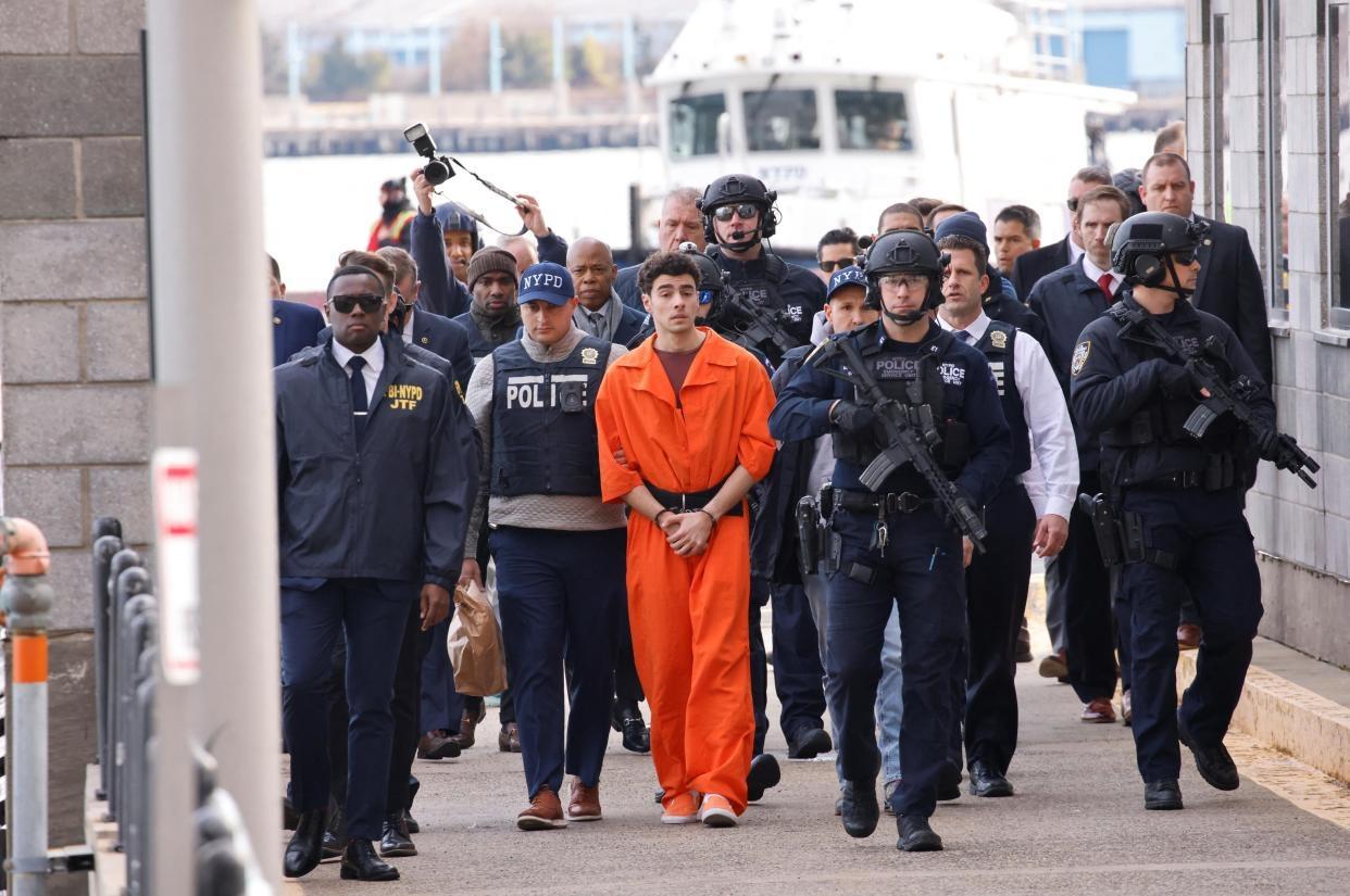 Luigi Mangione walking while handcuffed in an orange jumpsuit, with an absurd number of cops escorting him on either side
