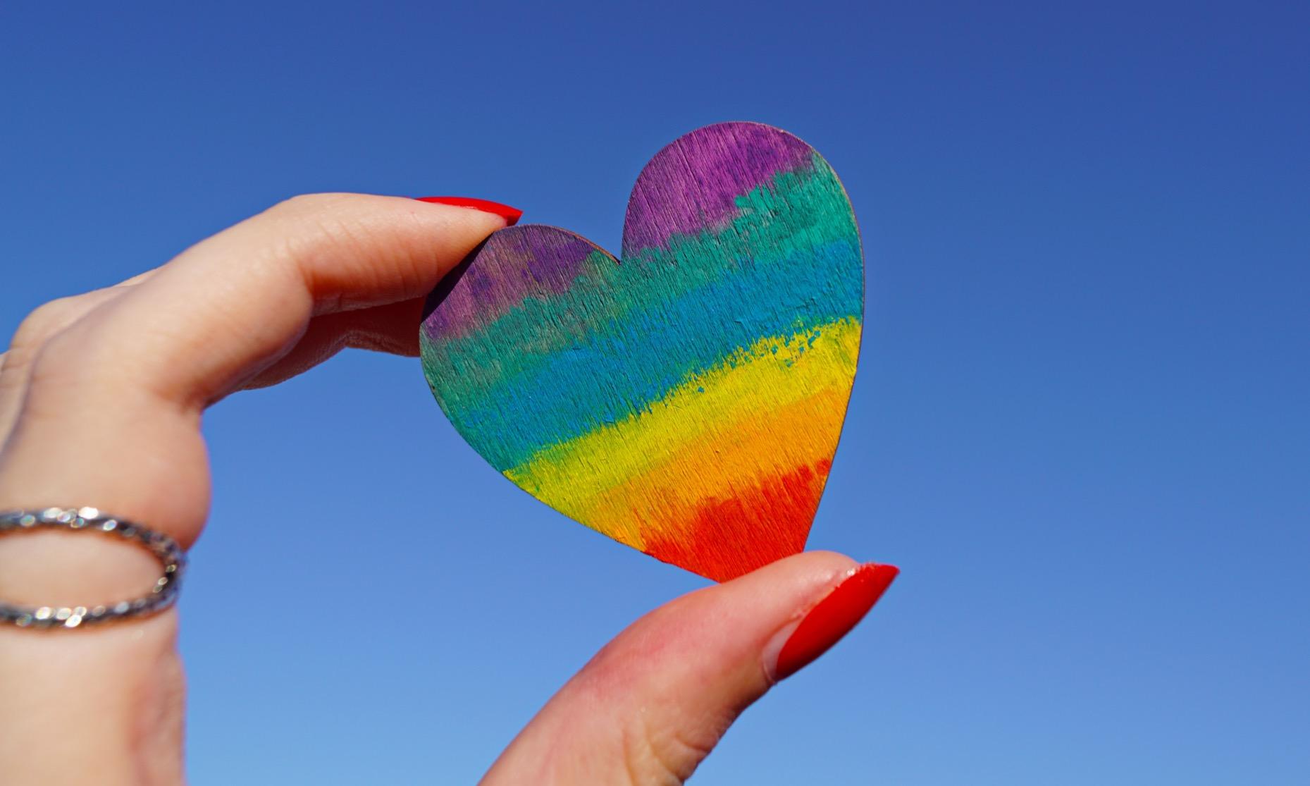 A person's hand with red nail polish holds a wooden heart painted with rainbow stripes against a bright blue sky. A silver ring is visible on their finger.