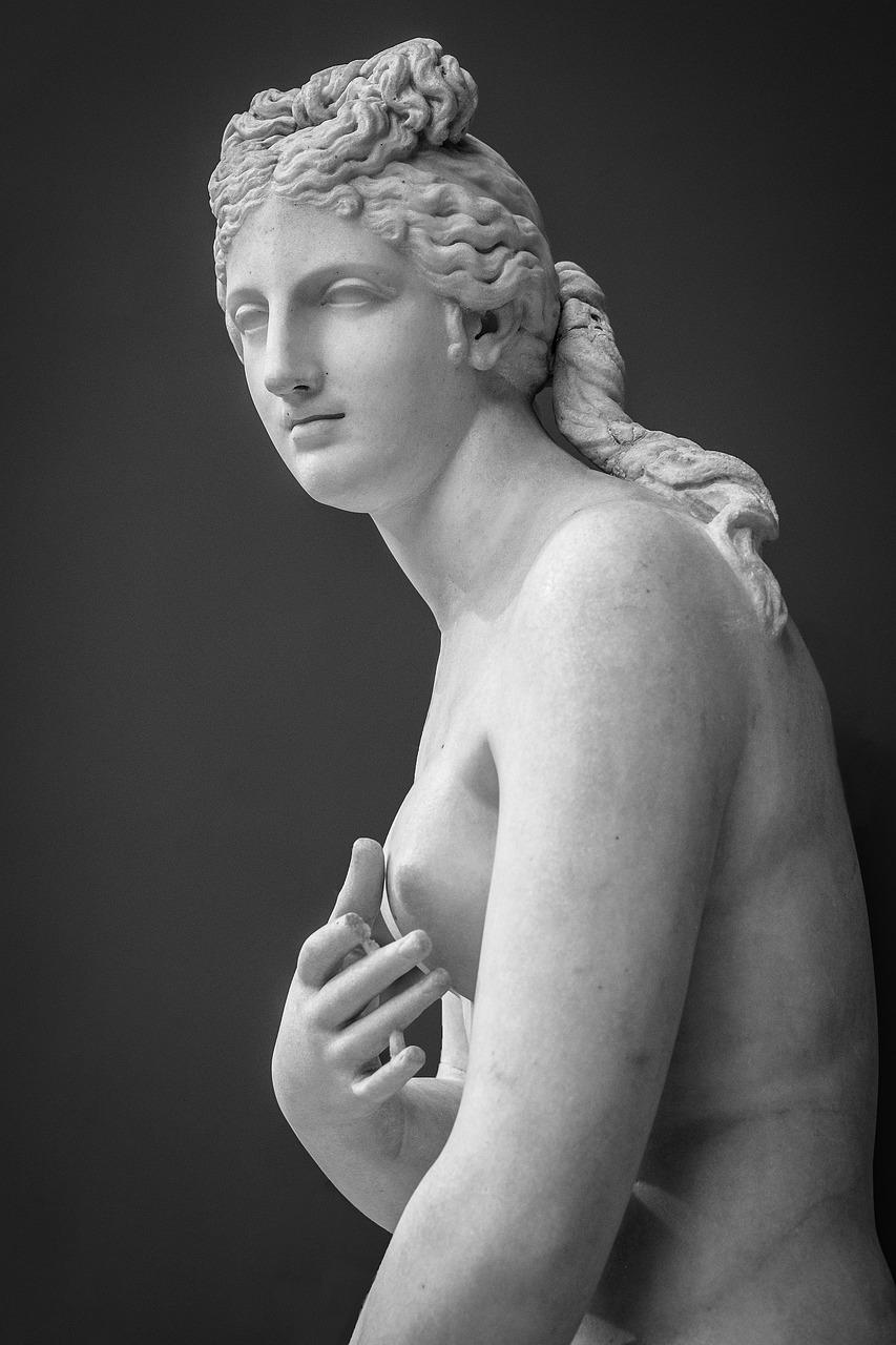 a classical marble statue of a nude female figure, Goddess Venus. The statue is depicted in a three-quarter view, with the figure's body turned slightly to the side, showcasing the graceful curves and musculature of the human form. The hair is intricately carved, with curls and waves that add texture and depth to the sculpture. The figure's left hand is gently placed over the chest, with fingers delicately positioned, while the right arm is relaxed at the side. The background is a solid, dark color, which contrasts with the light-colored marble, highlighting the details of the sculpture. The overall composition emphasizes the beauty and elegance of the human form, characteristic of classical art