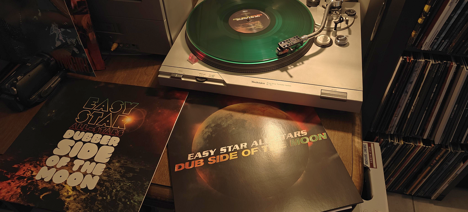 This is a cozy, warm-toned indoor photo focused on a record player and vinyl albums.

In the center, a silver Technics turntable is playing a translucent, emerald-green vinyl record. The tonearm is lowered onto the spinning disc.

In the foreground, propped up against the turntable, are two album covers from the "Easy Star All-Stars." The one on the right, titled "Dub Side of the Moon," features artwork of a large, cratered moon against a starry, orange nebula. To its left is a companion album titled "Dubber Side of the Moon," also with a space-themed cover.

The background is filled with shelves packed with a large collection of vinyl records, suggesting a dedicated music enthusiast's space.