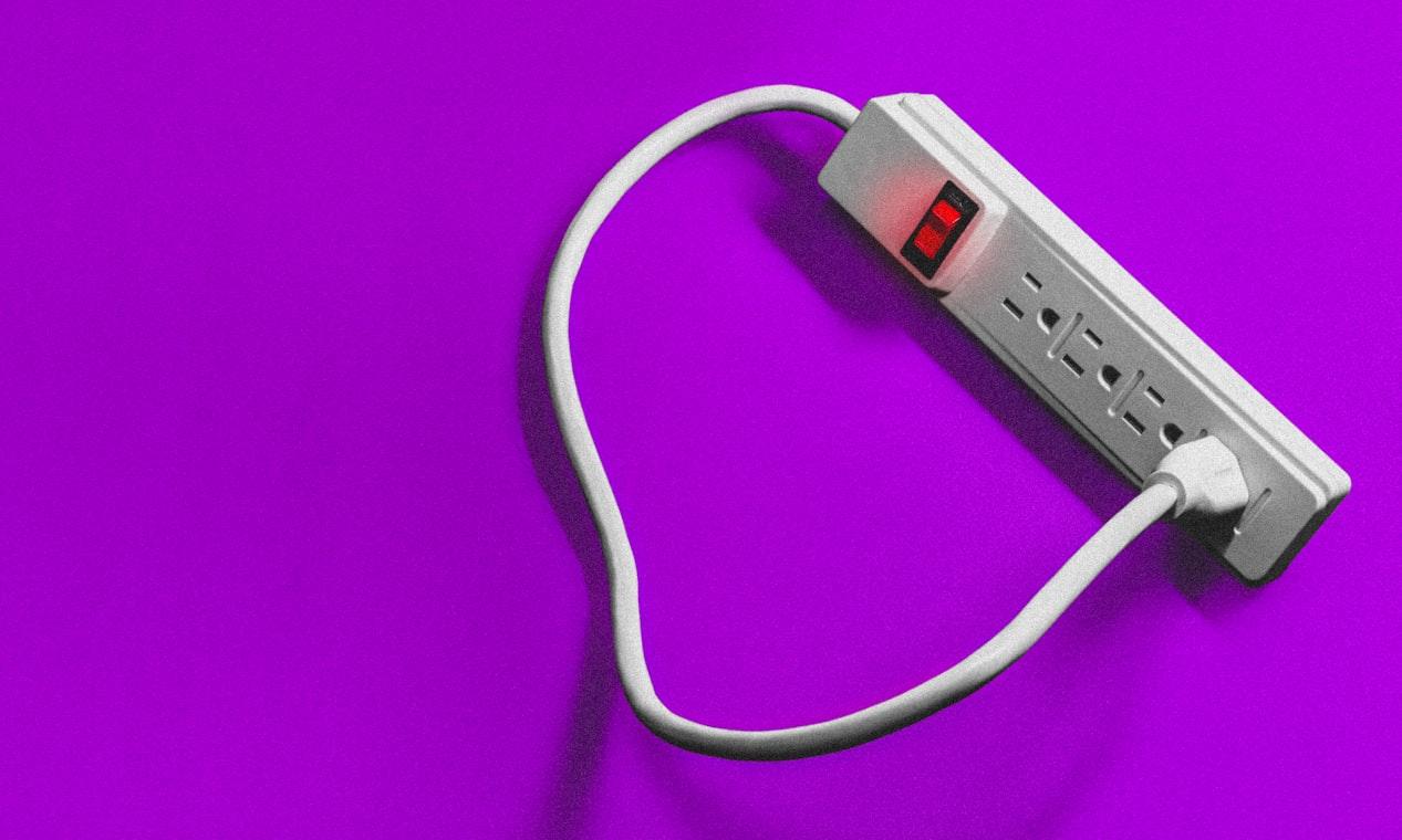 This is a studio-style photograph of a white power strip lying on a vibrant, solid purple background. The power strip has a red, illuminated "on" switch at the top. The power strip's own white cord is plugged into one of its own sockets, creating a closed <br />loop. The object is lit from the side, casting a soft shadow onto the purple surface.