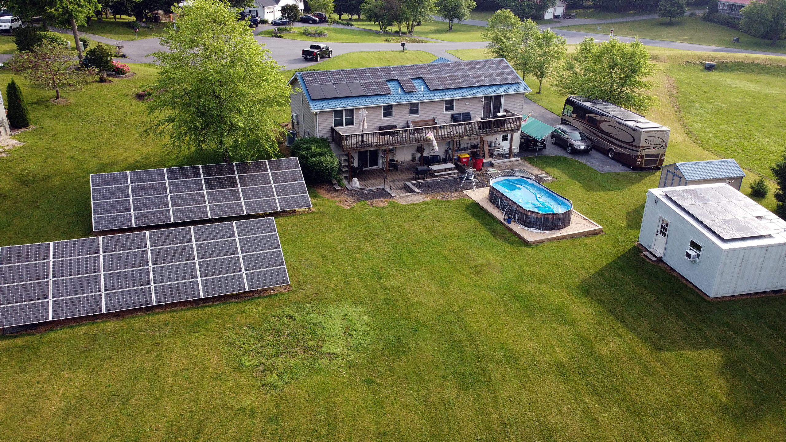 This is a high-angle, aerial view of a two-story house in a large, grassy yard. The property features extensive solar power, with two large, rectangular solar panel arrays mounted on the ground in the backyard. Additional solar panels are also installed on the house's roof and on a workshop. The backyard also contains an above-ground pool, and a large RV is parked in the driveway with solar panels on it.