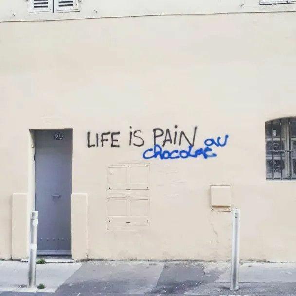 The image features a piece of street art where the phrase "LIFE IS PAIN" is modified with the French words "au chocolat