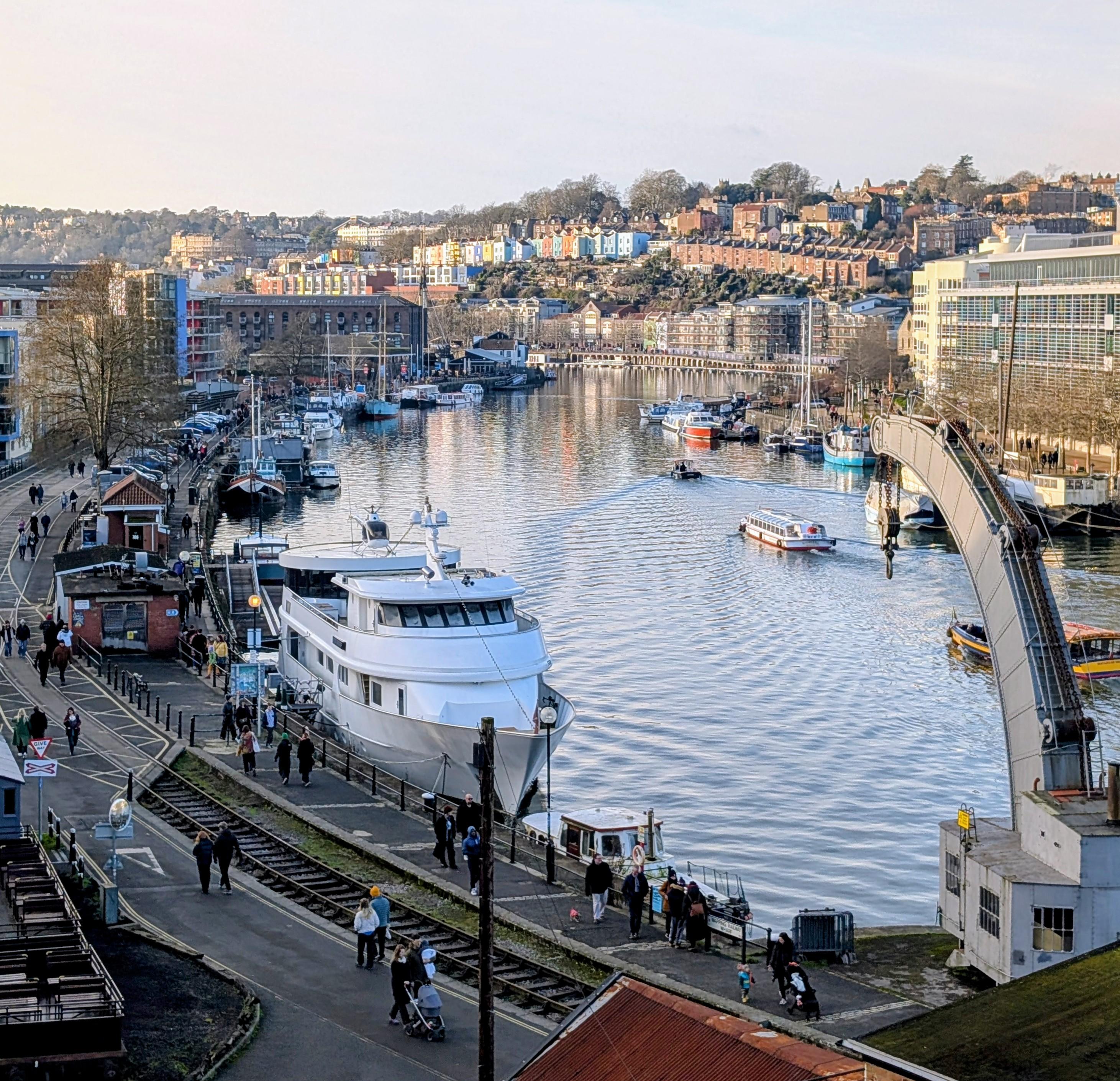 A view over a harbour with boats moored on the sides and several boats driving through. Sidewalks full of people and an old industrial railway. Beyond are colorful painted row houses along cliffs. The sky is light blue and a bit hazy.