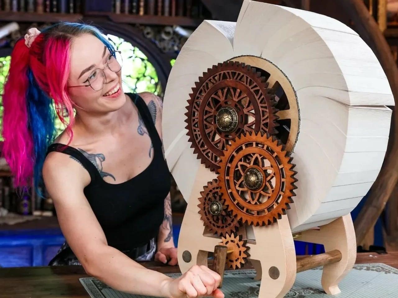 A photograph of a woman wearing glasses and pink tail in a ponytail hand cranking a 2 foot book wheel with complex cogs as its mechanicism