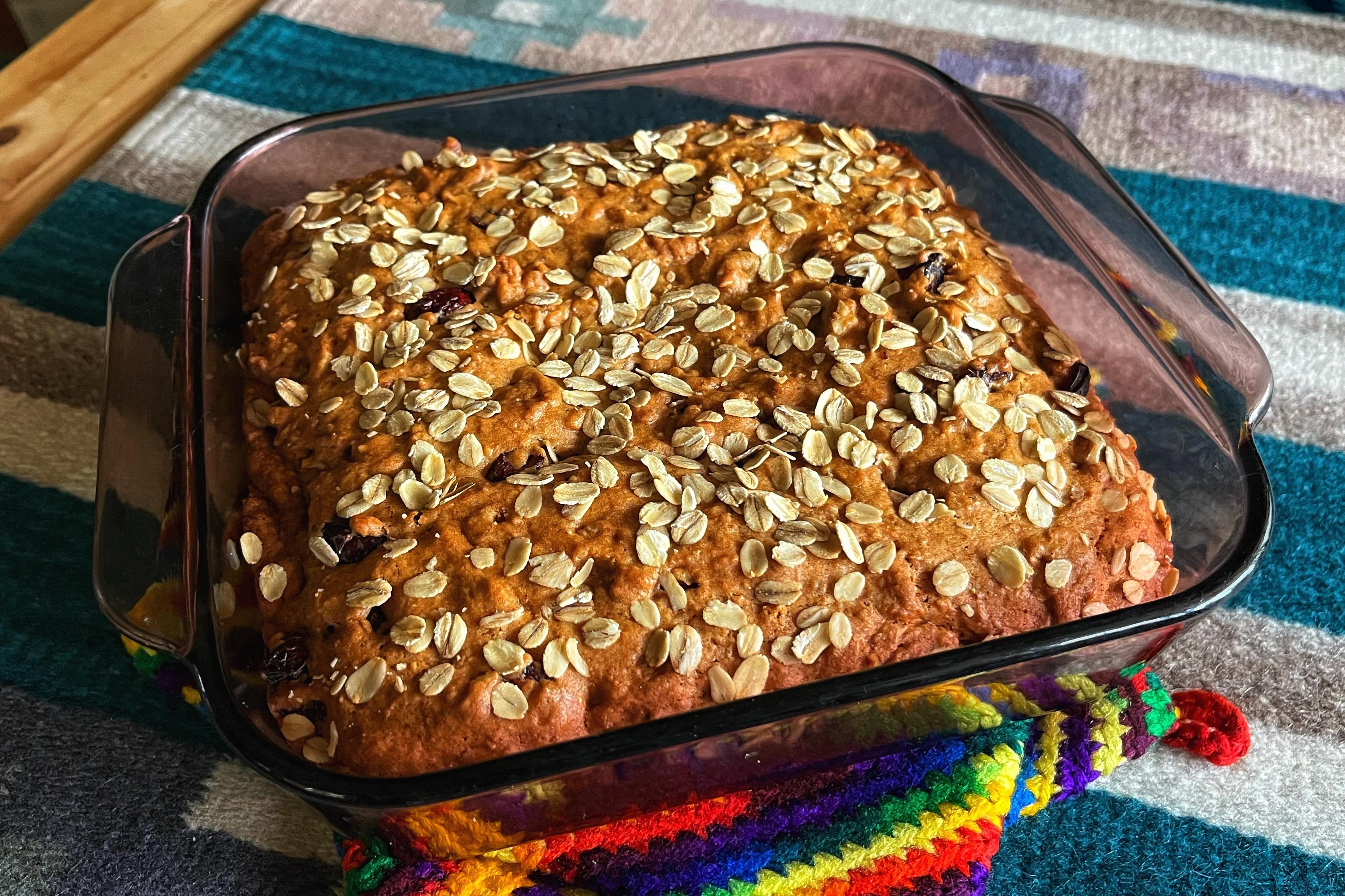 A square baking dish resting on rainbow colored potholders which are sitting on a table with a striped woven covering. The baking dish contains a freshly baked, golden brown applesauce cake with cranberries and walnuts, sprinkled with oats on top.