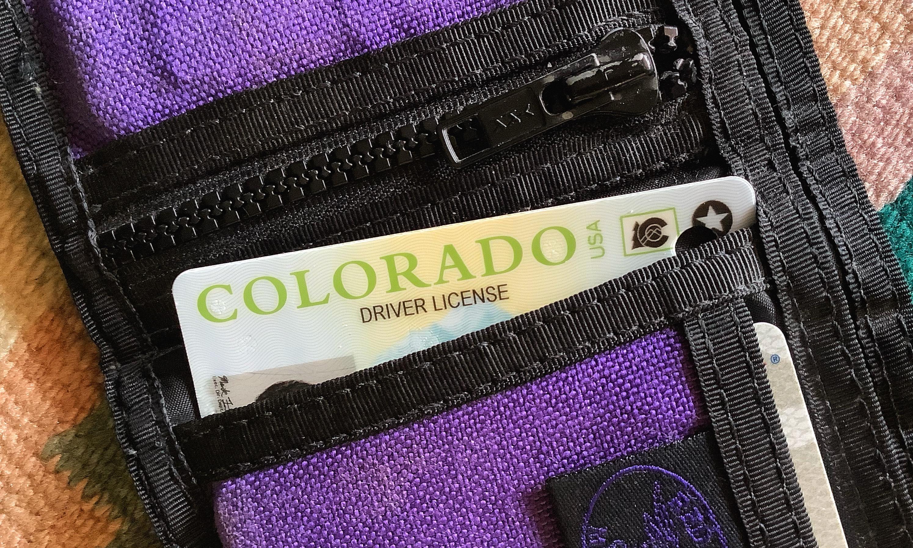 The top of a driver license card peeking out of a slot in a foldable, purple and black canvas wallet. The card says Colorado Driver License across the top.