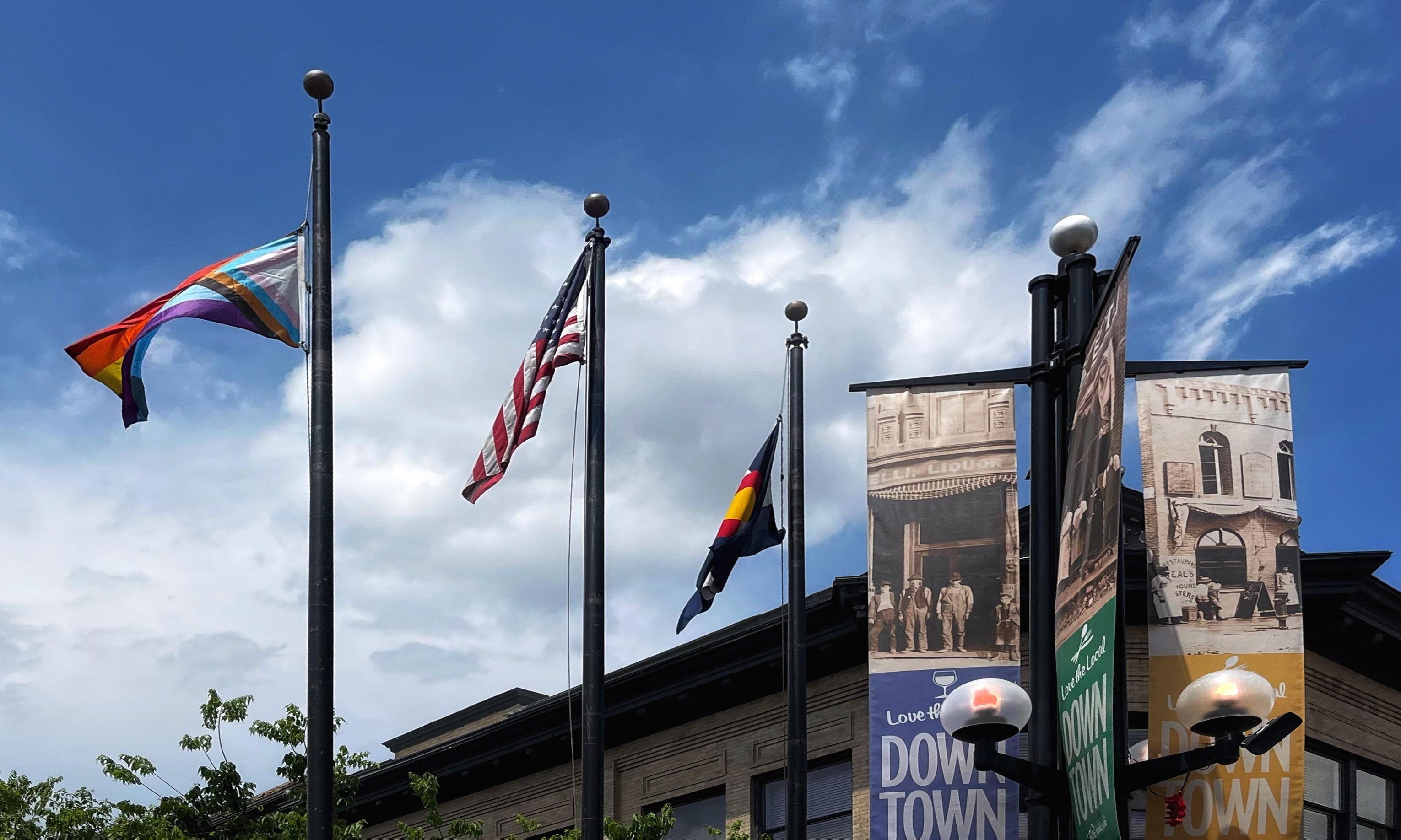 Looking upward at three, tall, side-by-side flagpoles, each with a different flag (left to right): a freshly raised Progress Pride flag for Pride Month, a US flag, and a Colorado flag. The flags are rustling in the breeze against a partly cloudy, midday background sky. There is another pole/lamppost to the right with globe lights and banners advertising Downtown on them, with the roofline of a brick building in the background.