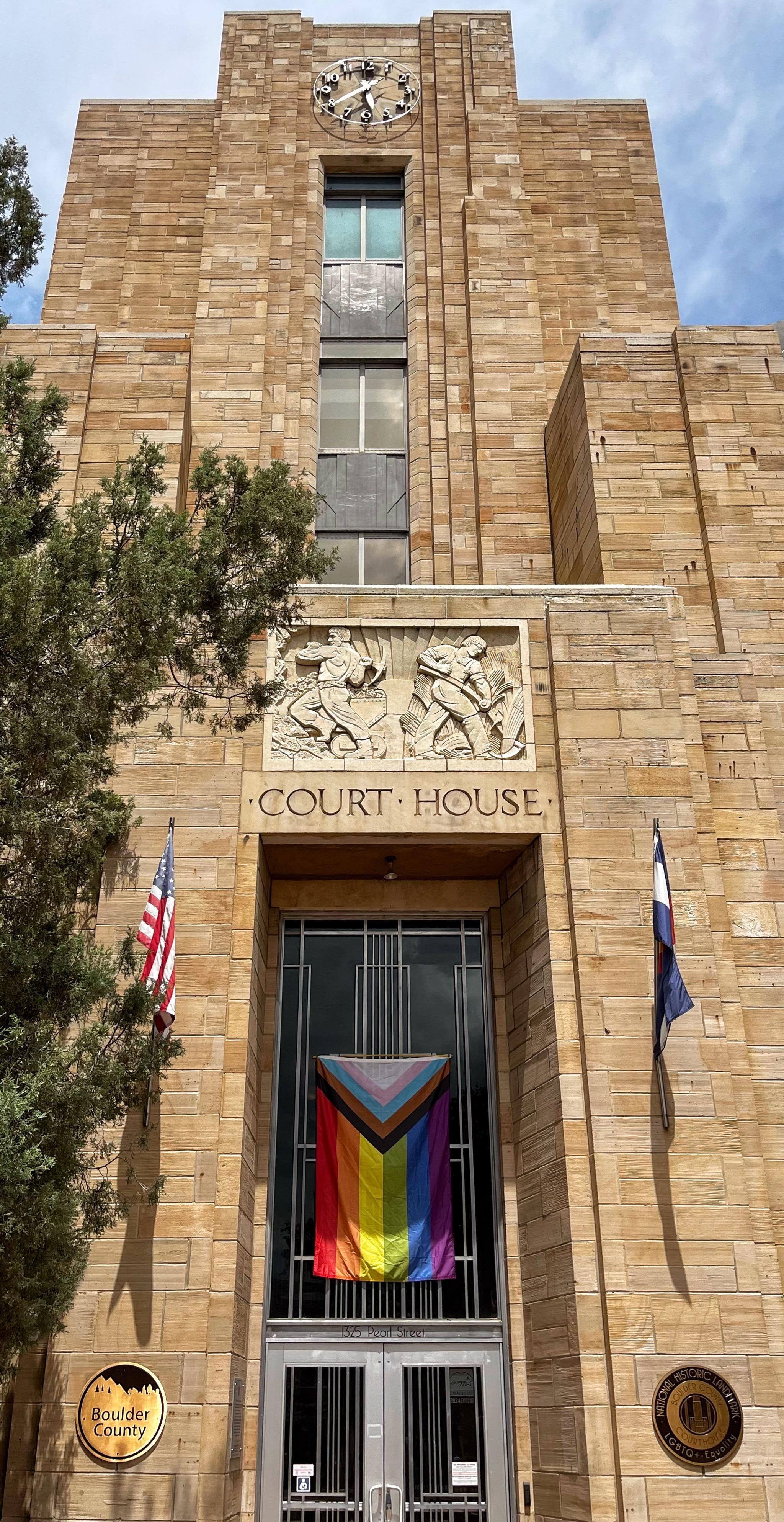 The front facade of the downtown, art deco style courthouse building constructed of tan sandstone with a clock tower at the top (with a non-working clock), a bas relief sculpture of men working in the center of the facade, plus the words Court House. There is a large Progress Pride flag hanging vertically above the entrance at ground level, flanked by a US flag to the left of the entrance doors and a Colorado flag to the right of the entrance doors.