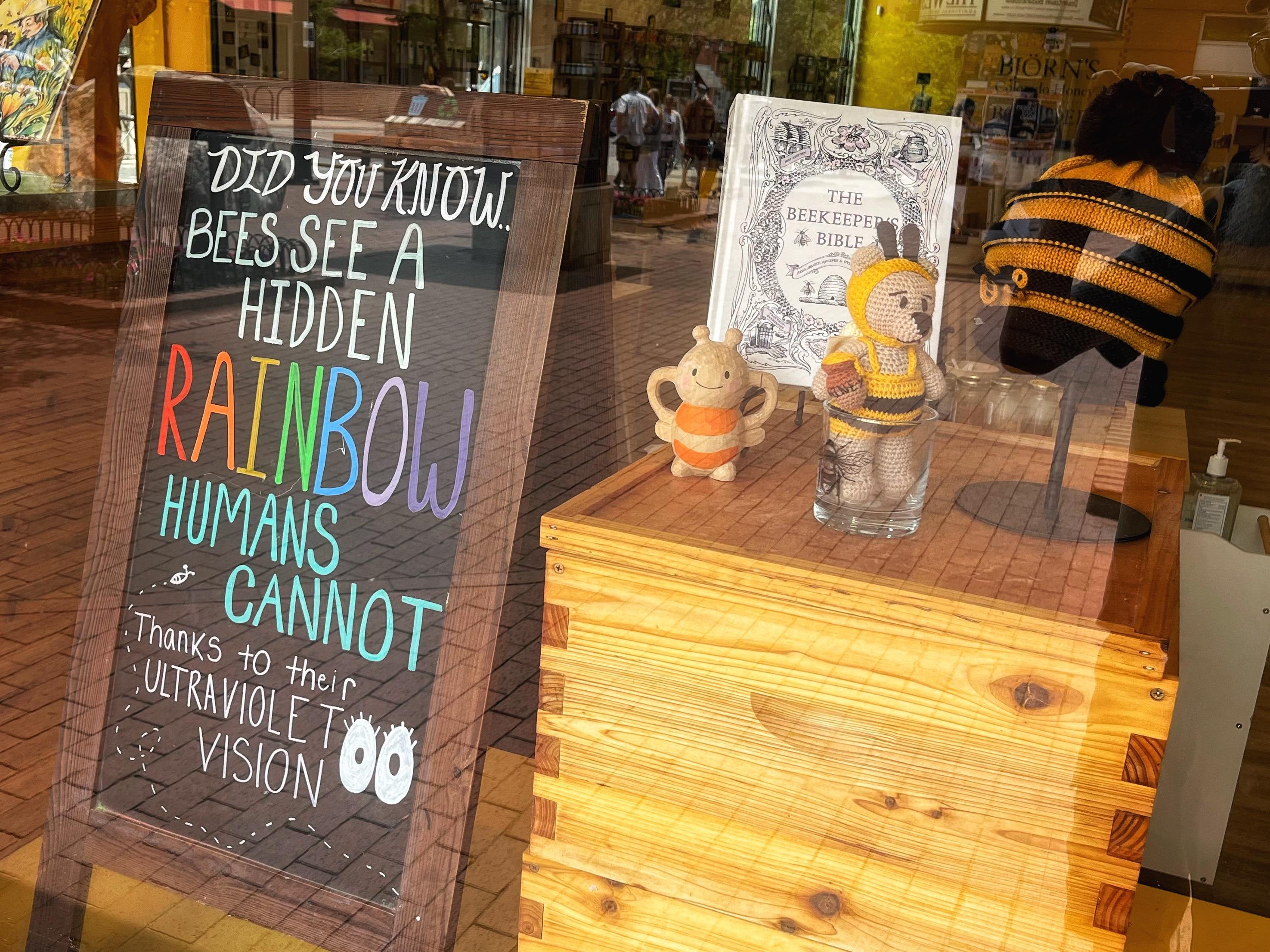 A window display at a local downtown honey store, with a signboard on the left that says, “Did you know bees see a hidden rainbow humans cannot, thanks to their ultraviolet vision?” On the right is wooden box with bee and honey-bear figurines on top, along with a beekeeping bible.