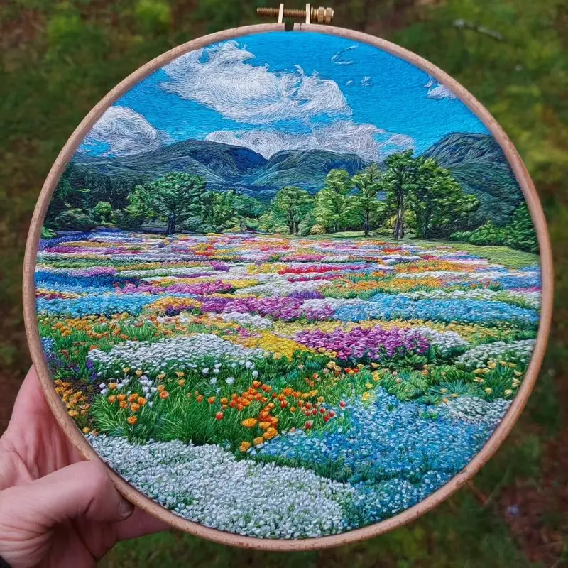 A, hand embroidery hoop art featuring a highly detailed, textured landscape of a vibrant wildflower field in green, blue, white and pink with green trees in the background before a darker green hill  range under a sunny bright blue sky