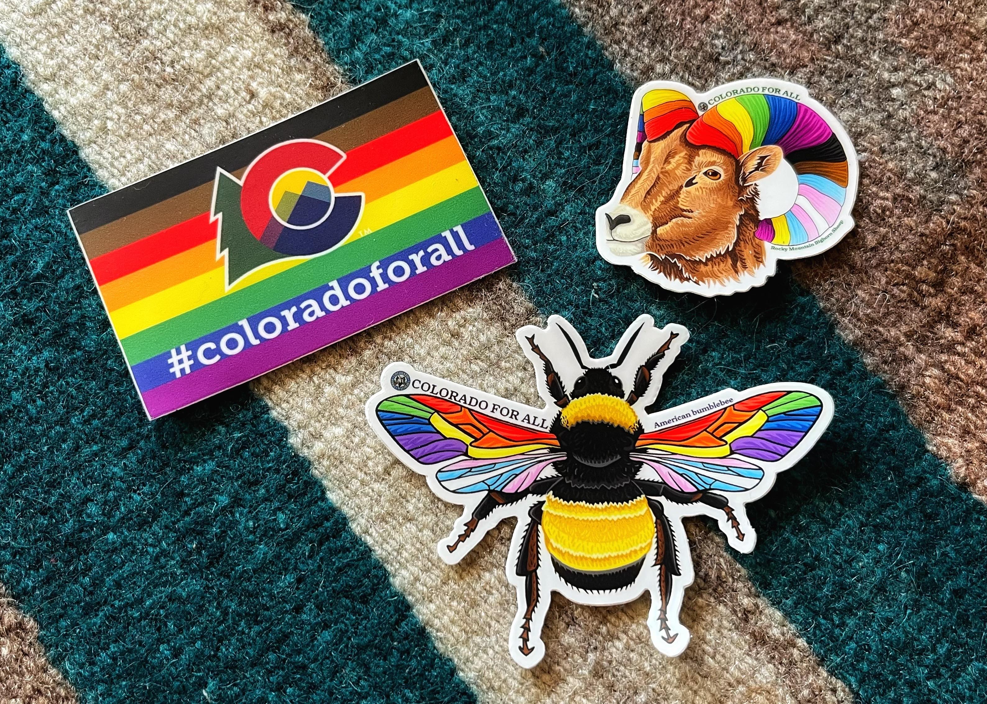 Three stickers on a table from Colorado Parks and Wildlife on a table: 
— a state flag with horizontal stripes in the progress pride colors (brown, black, red, orange, yellow, green, blue, purple), the stylizedColorado state logo, and the hashtag text #coloradoforall
— a Rocky Mountain bighorn sheep with horns spiraling behind its head that are colored with all the different pride colors
— a black and yellow striped American bumblebee with wings colored in all the different pride colors