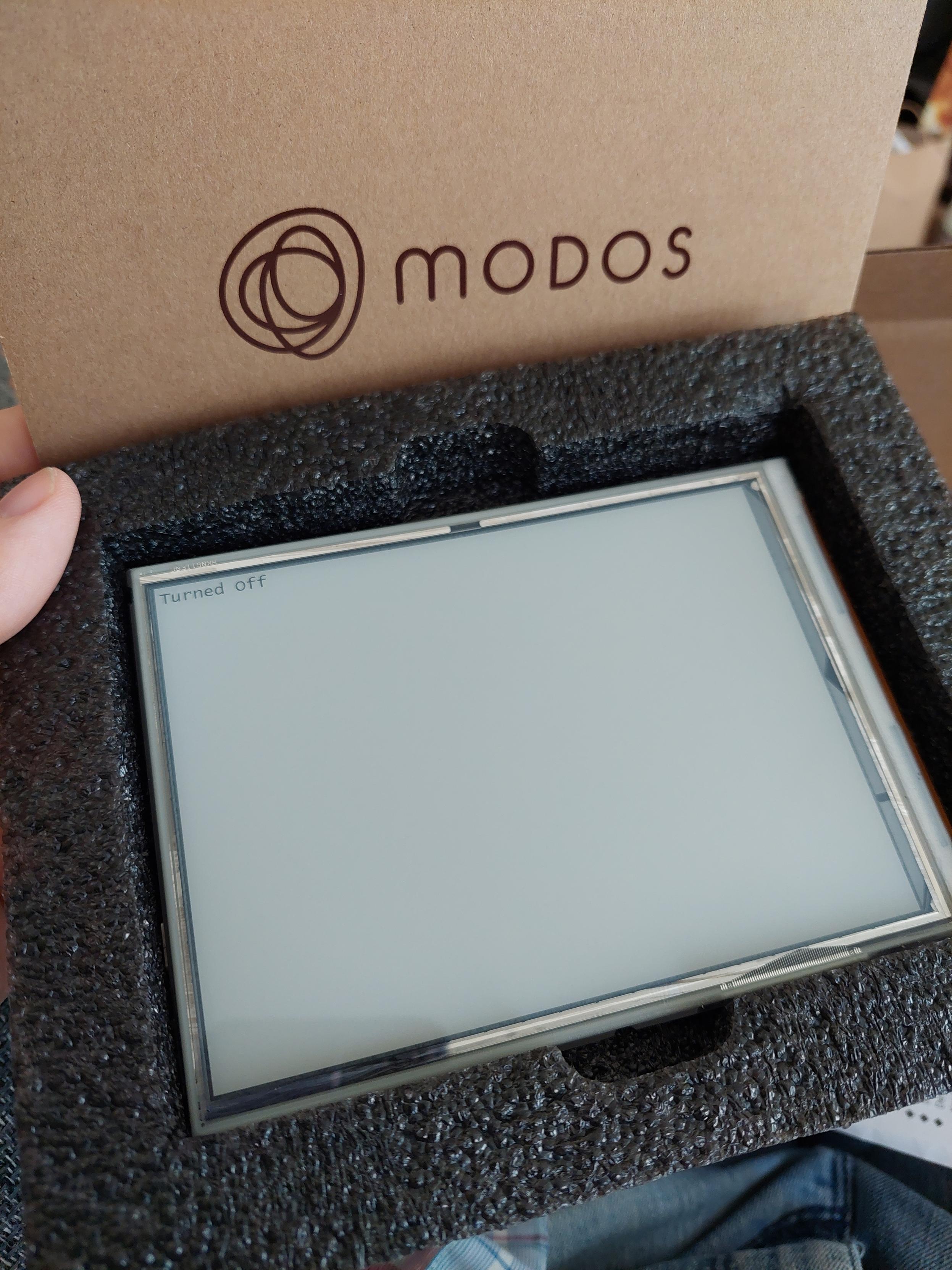 Photo of a 4x3 e-ink display in a foam liner. The name modos is branded on the box.