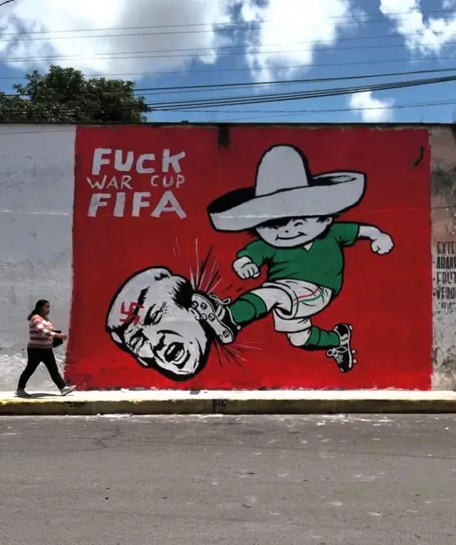 A mural of acartoon boy representing Mexico, dressed in a green national football kit and wearing a traditional sombrero. He is kicking the severed head of Trump, which  has a red swastika on the forehead.

The title is "FUCK WAR CUP FIFA", against a vibrant red background.