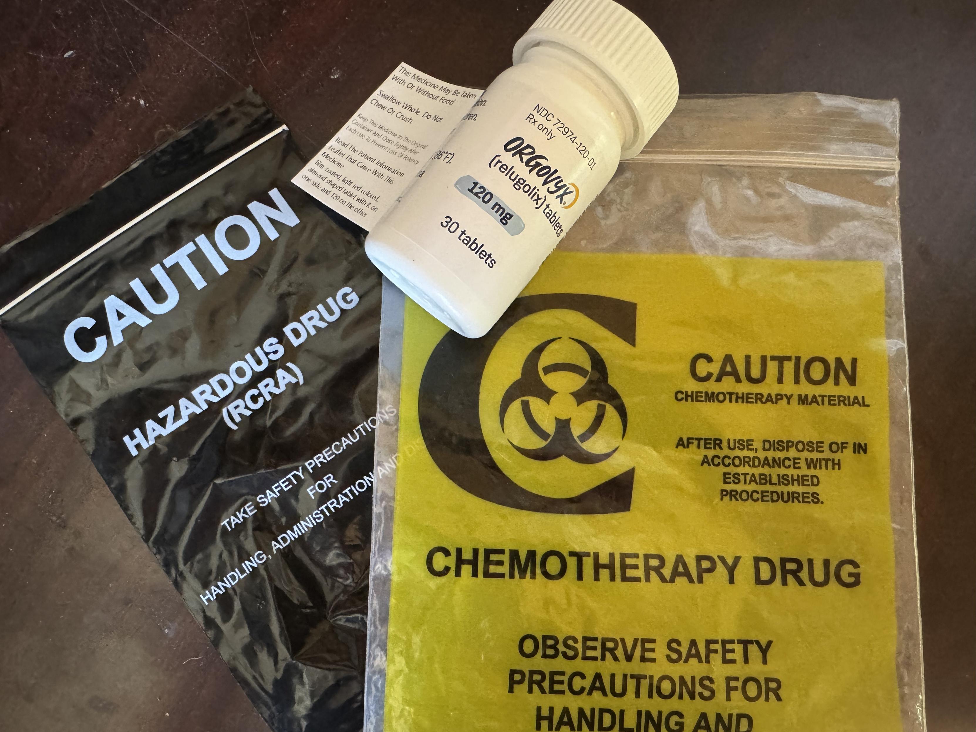 A bottle of the prostate cancer drug Orgovyx jauntily staged over plastic chemotherapy bags plastered with warnings about how hazadrous they are.