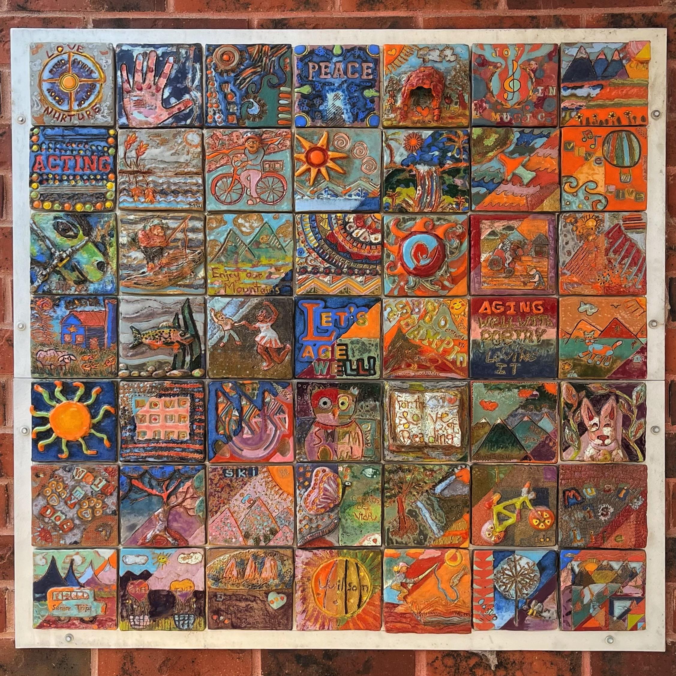 A mosaic of 49 colorful, handmade, ceramic tiles arranged in a 7x7 grid and mounted on a red brick wall outside a public building. Each tile tells a little story about aging well and being active. There are images and text of things like bicycles, sunshine, mountains, animals, flowers, birds, fish, sports, encouraging affirmations, and more etched and painted into each wonderful, unique tile.
