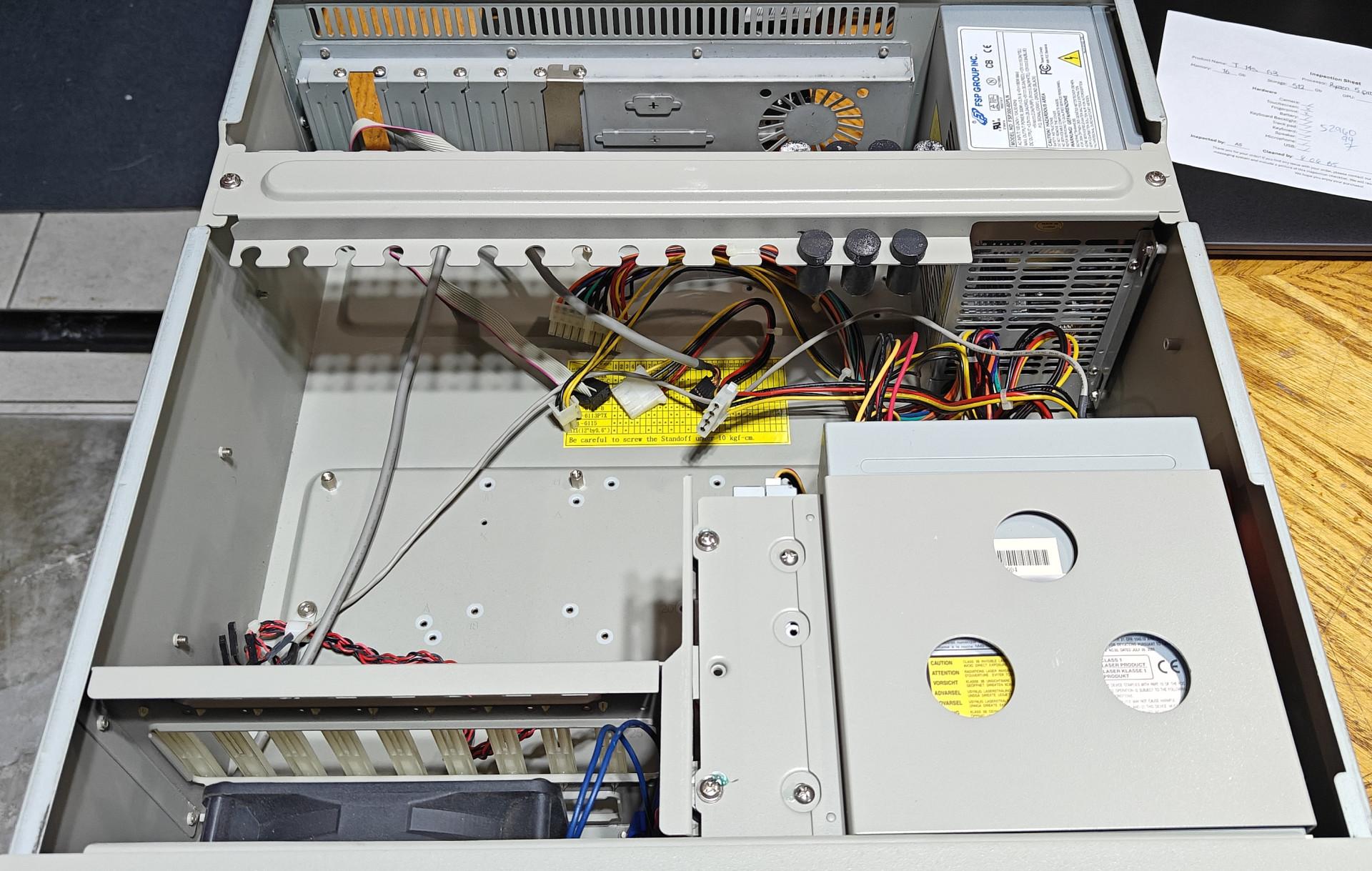 An overhead view reveals the open interior of a beige industrial rackmount computer chassis. The motherboard tray is completely empty, showing pre-installed brass standoffs ready for a build, alongside a prominent yellow warning sticker about standoff screw torque. An integrated hold-down bracket with adjustable black rubber bumpers spans across the middle to secure full-length expansion cards. Loose, multi-colored internal power cables extend from an FSP Group power supply mounted in the rear corner. A large cooling fan sits at the front corner, and a metal drive bay housing a disk drive occupies the right side.