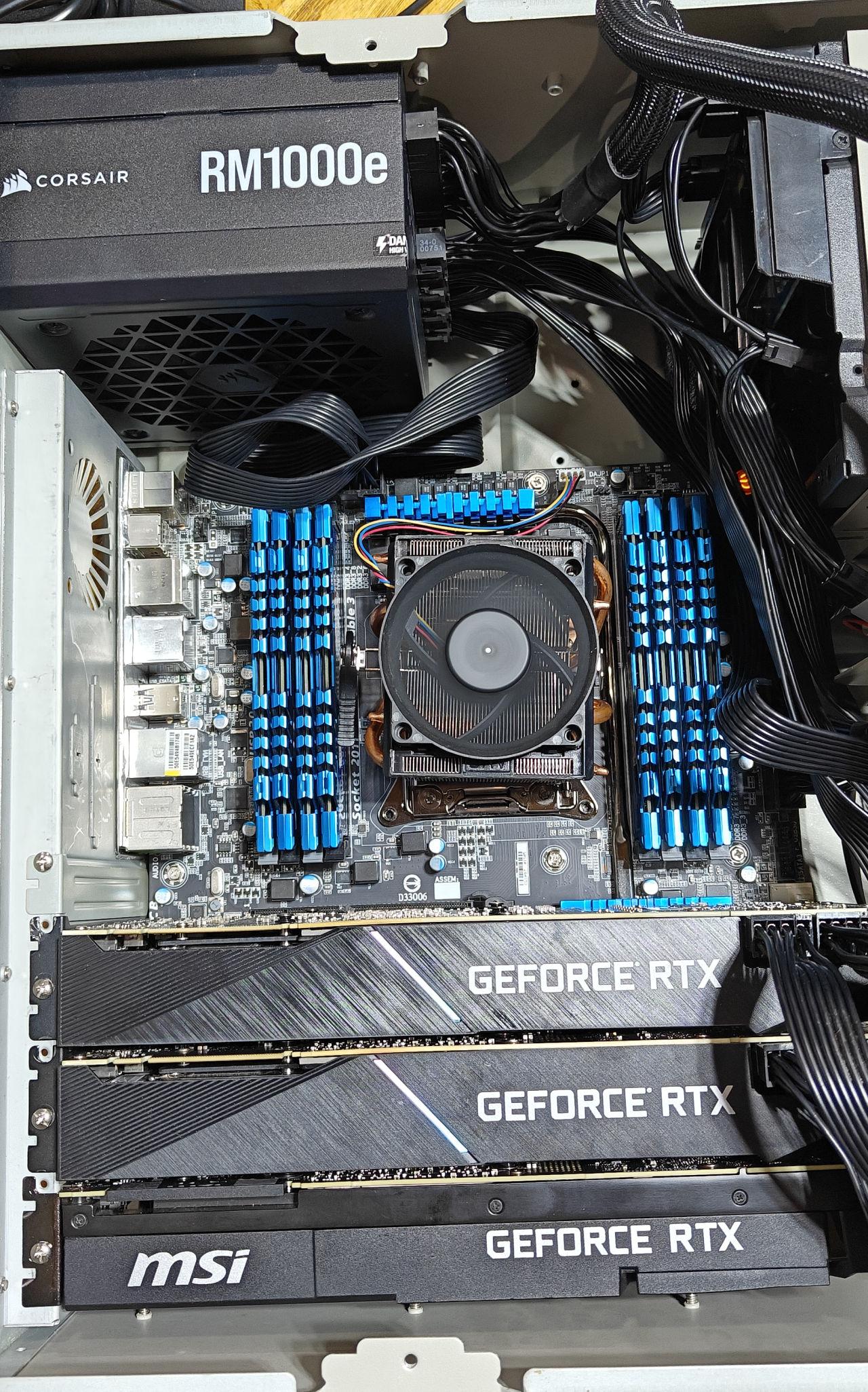 A direct overhead view looking down into the open metal computer chassis, detailing the internal hardware configuration. In the lower section, three massive Nvidia GeForce RTX graphics cards are installed closely together in parallel PCIe slots, with the bottom unit clearly showing a prominent white MSI logo. Heavy black power cables plug into the side of the top graphics card. In the center, a motherboard features an Intel Socket 2011 CPU interface equipped with a central copper heat pipe cooler and a black cooling fan. Flanking the processor cooler on both sides are eight memory modules fitted with striking metallic blue heat spreaders, filling every available DIMM slot. At the top left, a black Corsair RM1000e power supply is positioned with an array of black modular cables emerging from its side and routing across the chassis, while a tiny amber indicator light glows on the motherboard circuit board components nearby.
