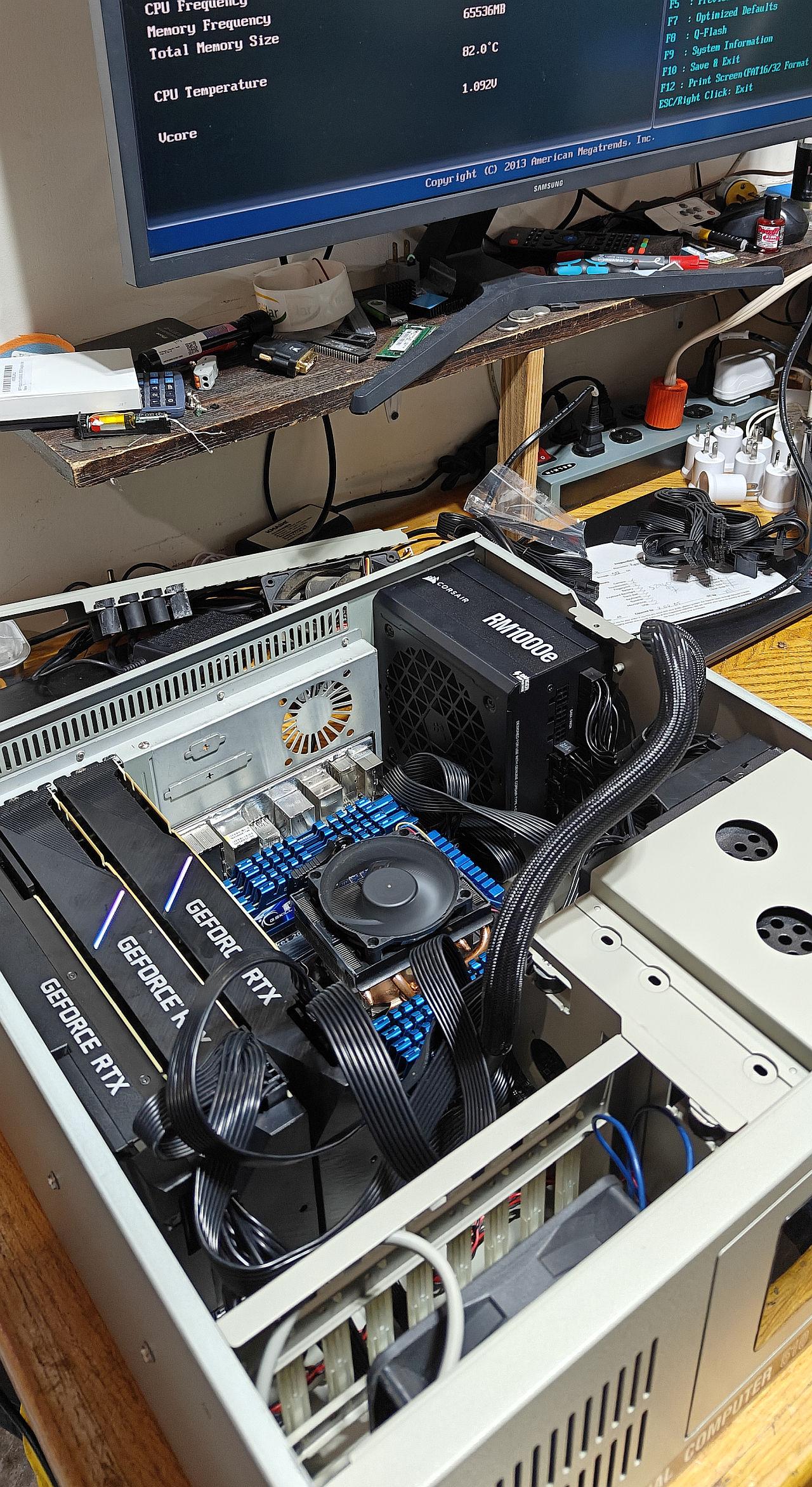 An angled top-down view shows an open computer chassis revealing a high-performance system built inside a vintage beige desktop case. Three large Nvidia GeForce RTX graphics cards are installed parallel to each other, taking up the lower left section. Above them, a motherboard features eight sticks of RAM with vibrant blue heatsinks flanking a central CPU cooler with a spinning black fan. A Corsair RM1000e modular power supply is mounted vertically near the upper right side, with numerous bundled black power cables routed throughout the metal interior. A thick, braided black sleeve wraps around another major cable bundle sweeping across the components toward the front frame. In the background, a monitor displays a BIOS setup screen showing sixty-five thousand five hundred thirty-six megabytes of system memory and a high CPU temperature of eighty-two degrees Celsius, resting on a cluttered wooden workstation shelf filled with tools and various small electronic components.