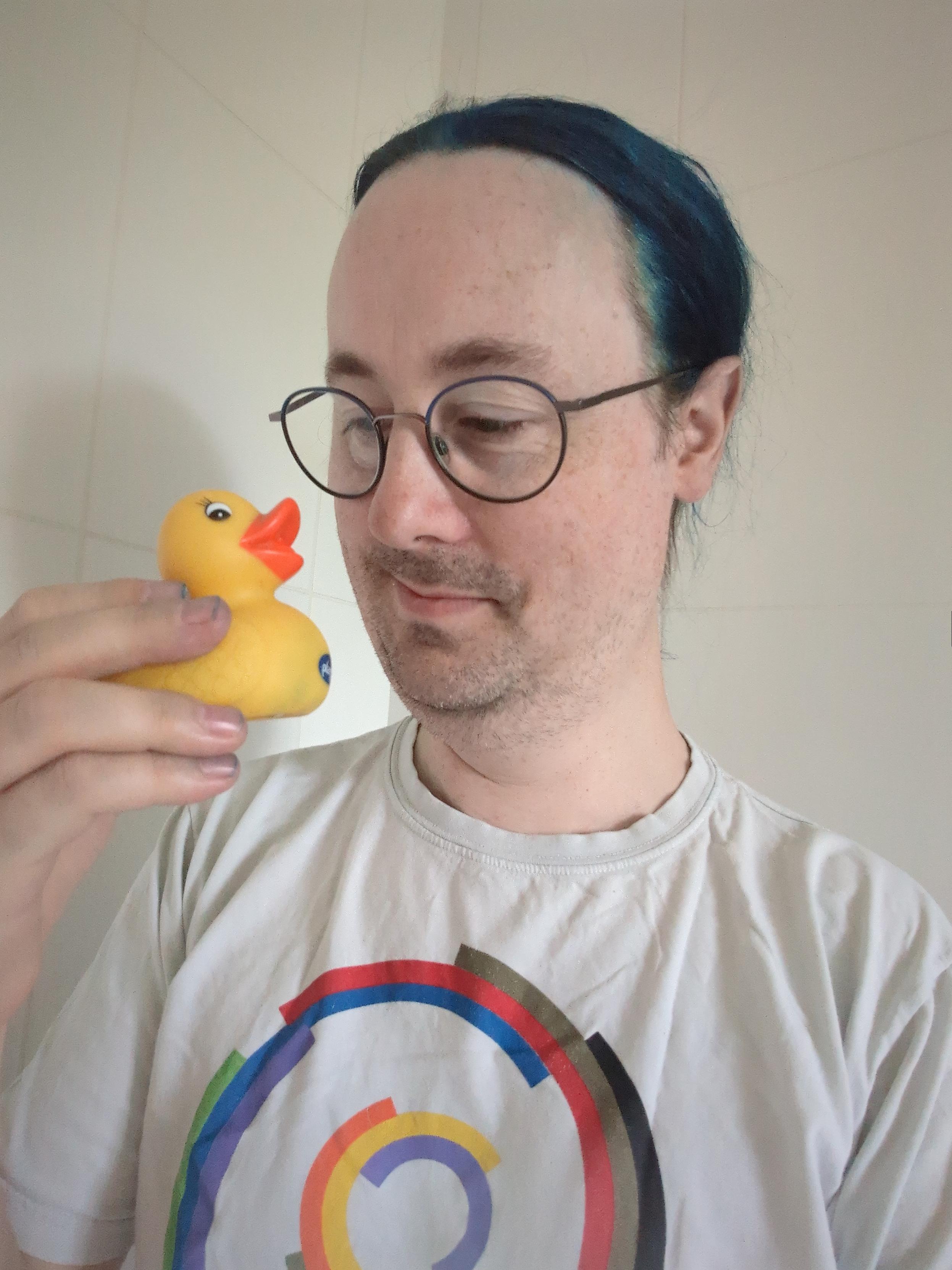 Me holding up and looking at a small yellow bath duck. I'm a white guy with medium-length blue hair, wearing glasses and a Debian Diversity T-shirt
