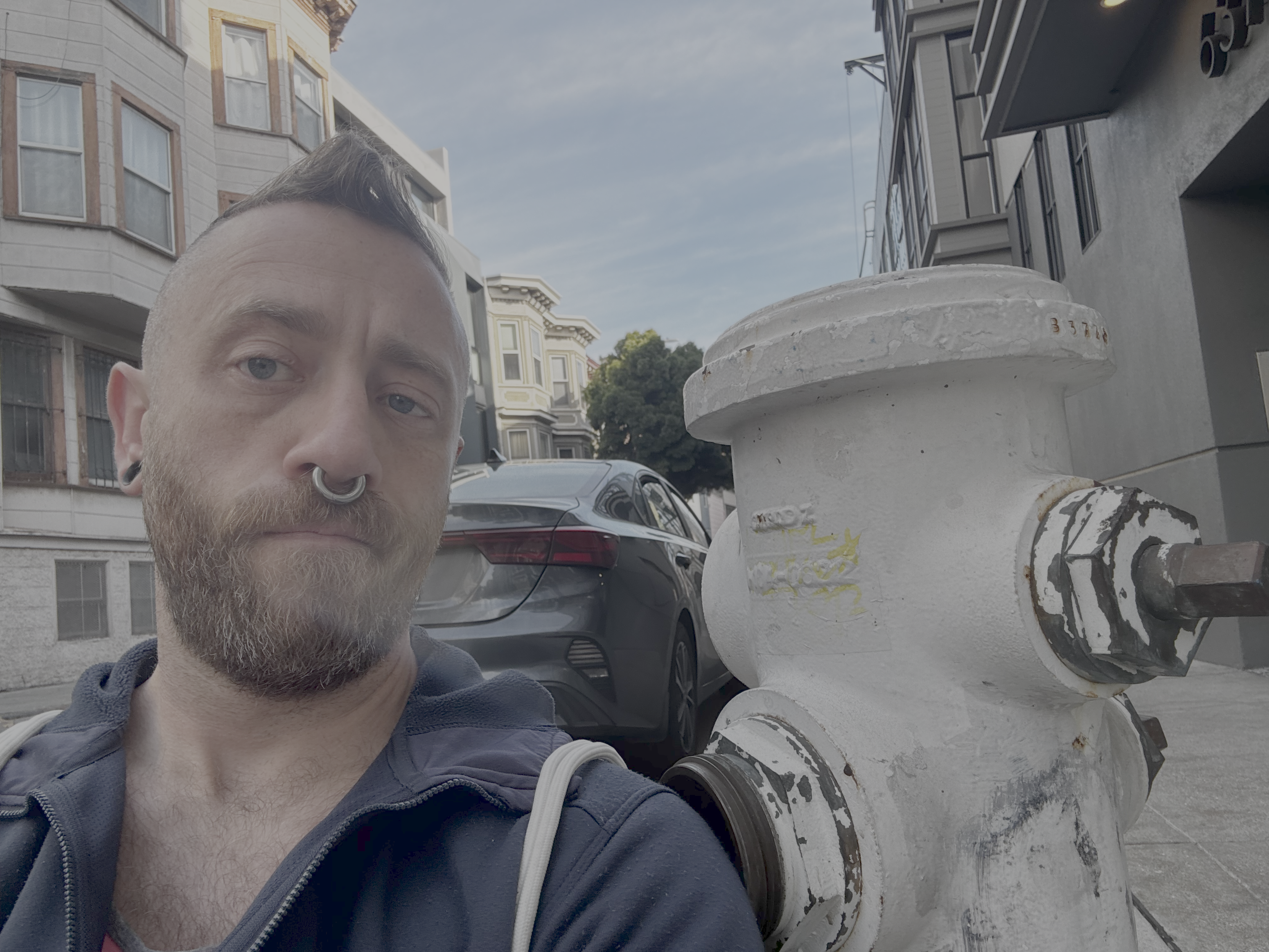 Bearded man with a septum ring, undercut, and blue eyes gives deadpan in a blue hoodie beside a weathered white fire hydrant. Foggy SF street. We are both structurally load-bearing.