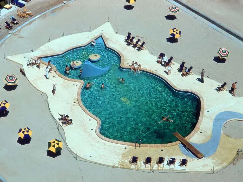 A large cat shaped swimming pool with turquoise water  surrounded by concrete and sun umbrellas