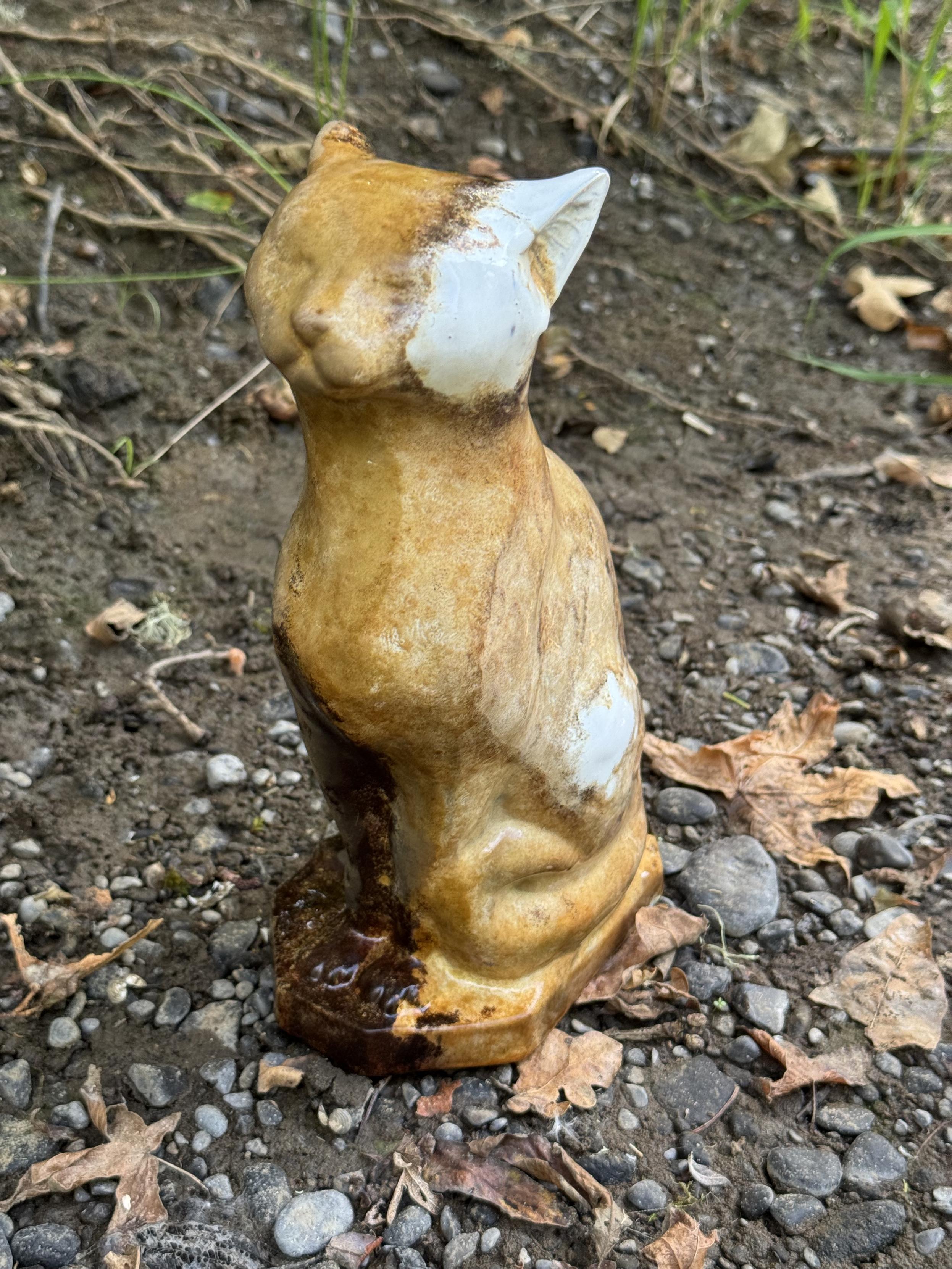 A stylized ceramic cat figurine about a foot tall. It used to be white, but is it now stained rust orange by the riverbed. Only its left ear, jowl, and bit of flank remain white.

It stands upright on a gravel bank strewn with rounded river pebbles and fallen dead leaves.

The cat stands tall as if stretching up for head scratches. It a self satisfied smirk and closed eyes. The tail is wrapped around to the front paws.