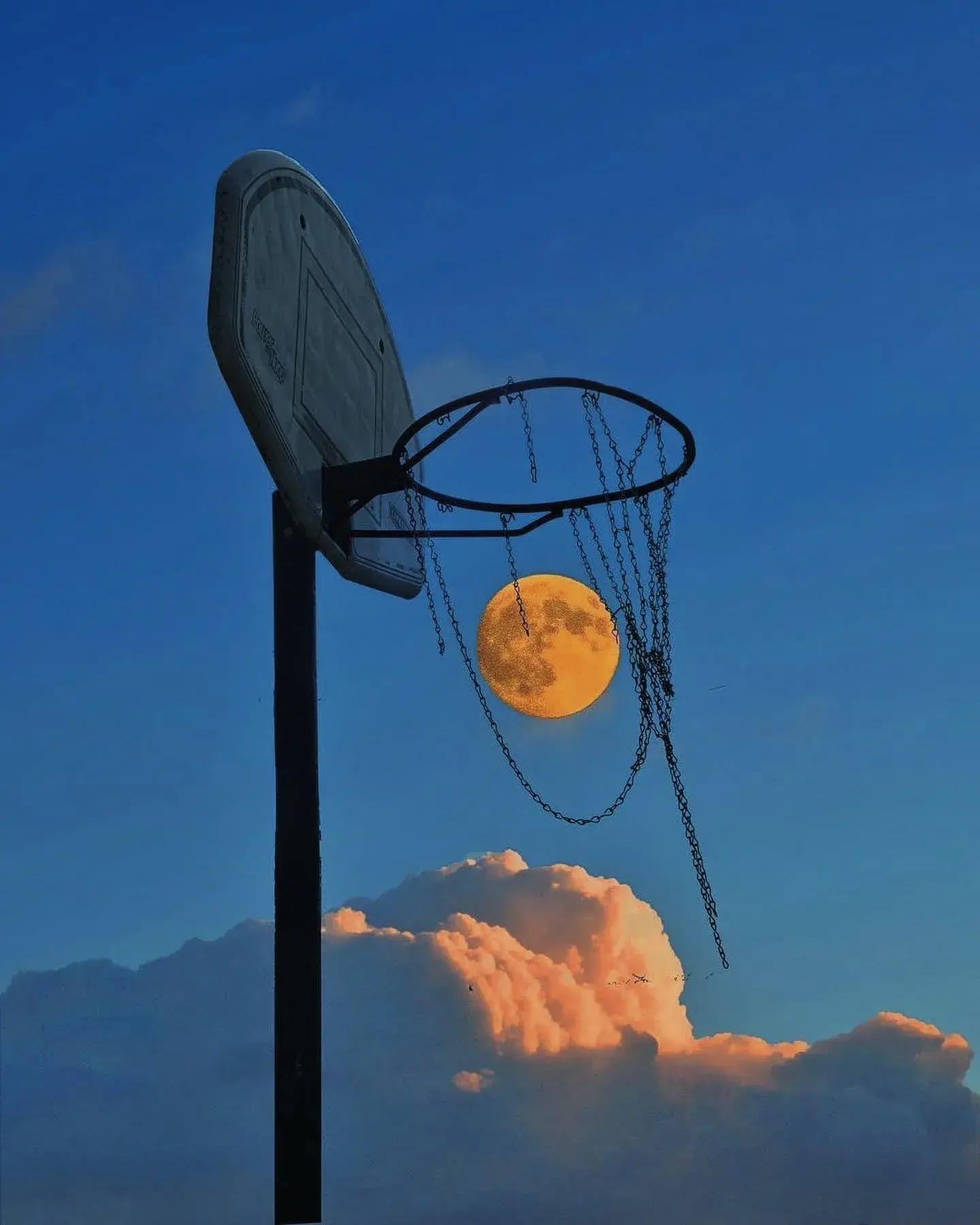 A photo of a basketball hoop set against a deep blue sky, except the moon is captured inside the netting