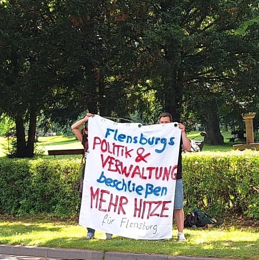 Zwei Menschen halten ein Banner auf dem steht "Flensburgs POLITIK & VERWALTUNG beschließen MEHR HITZE für Flensburg