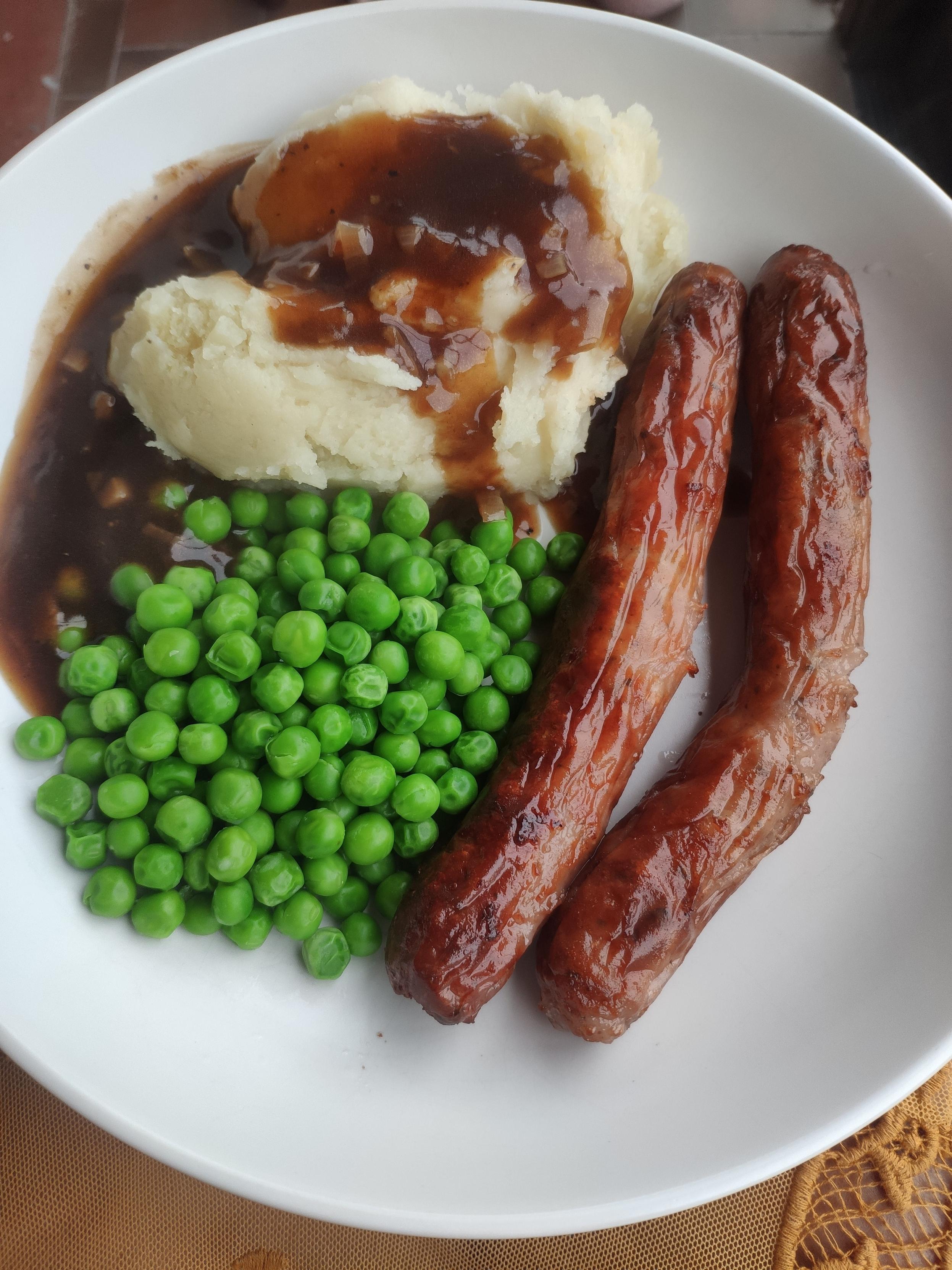 A white plate with two perfectly cooked sausages, mash, brown gravy and green peas.