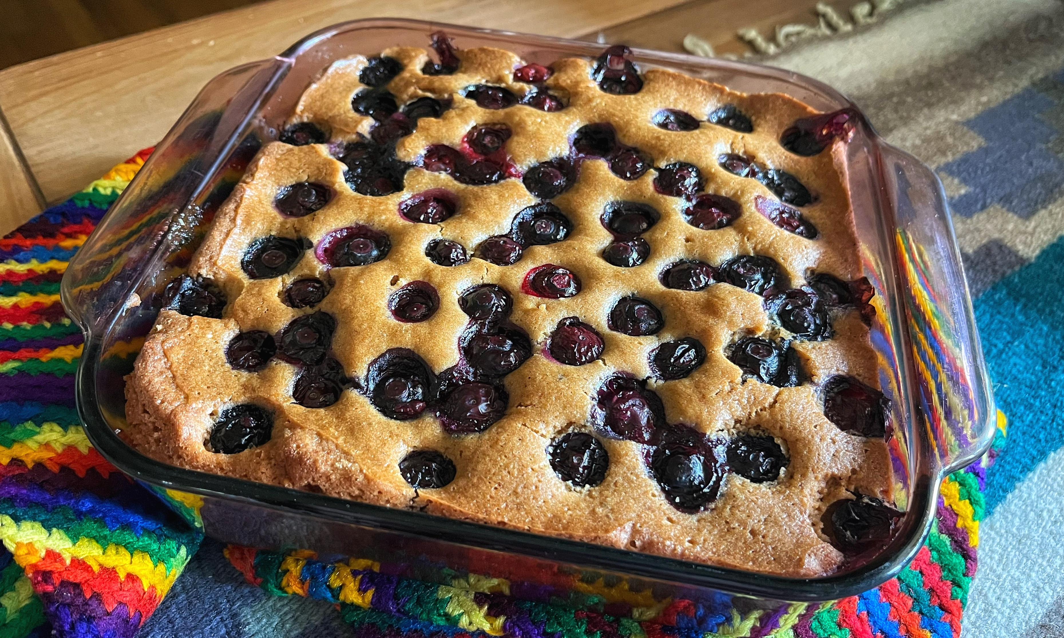 Glass baking just out of the oven and resting on rainbow potholders on a wooden table. The dish has a freshly baked upside-down cake in it, golden brown with blueberries baked into the top.