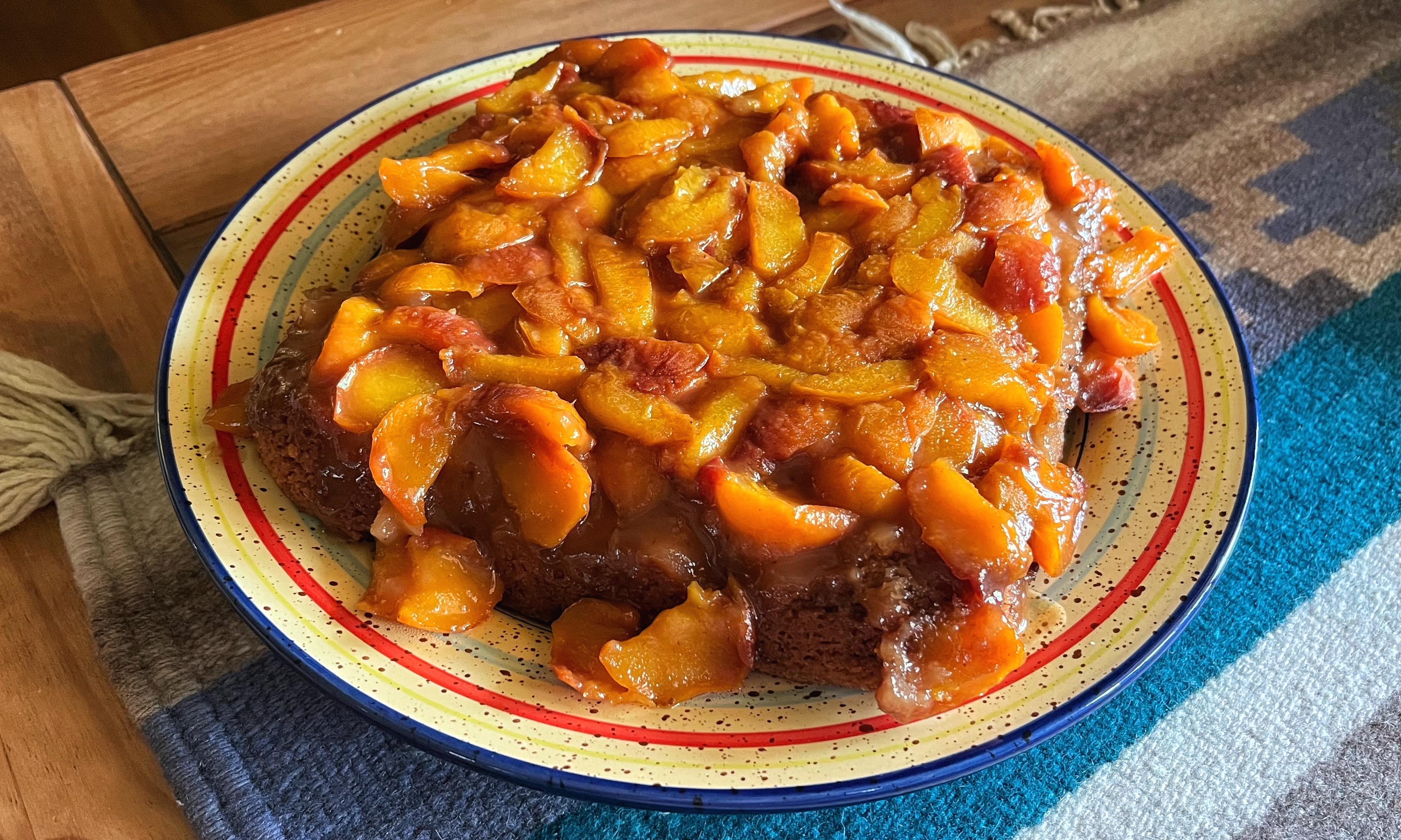The final presentation: A peach upside-down cake, now flipped onto a large, colorful, round platter and resting on a wooden table — the baked, golden-orange, glazed peaches that were on the bottom are now on top and are slowly oozing off the edges of the cake onto the platter.