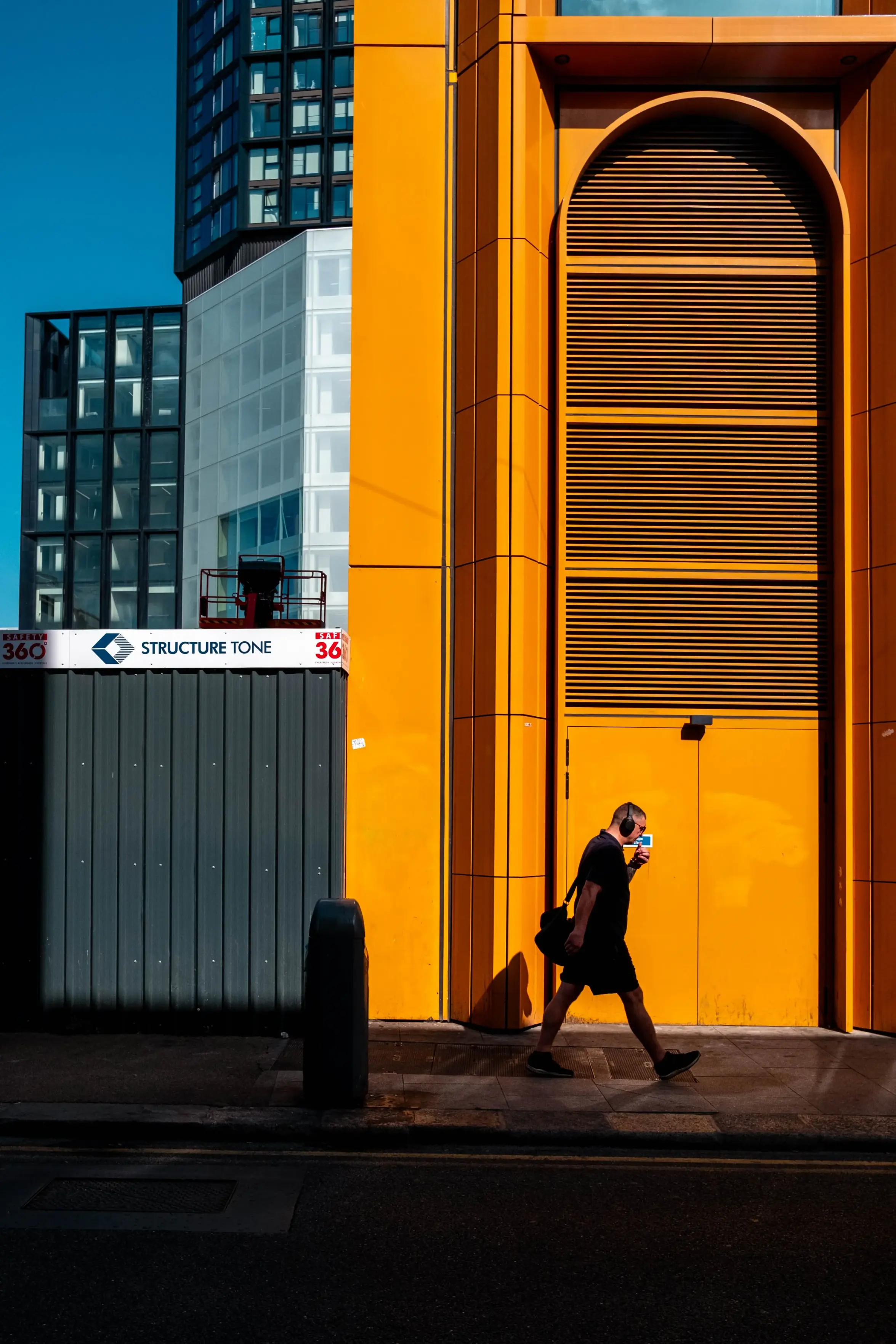 A photo of an urban street scene featuring a striking, vibrant yellow architectural facade with a tall arched entryway and louvered panels, alongside a pedestrian in silhouette wearing headphones and carrying a bag.