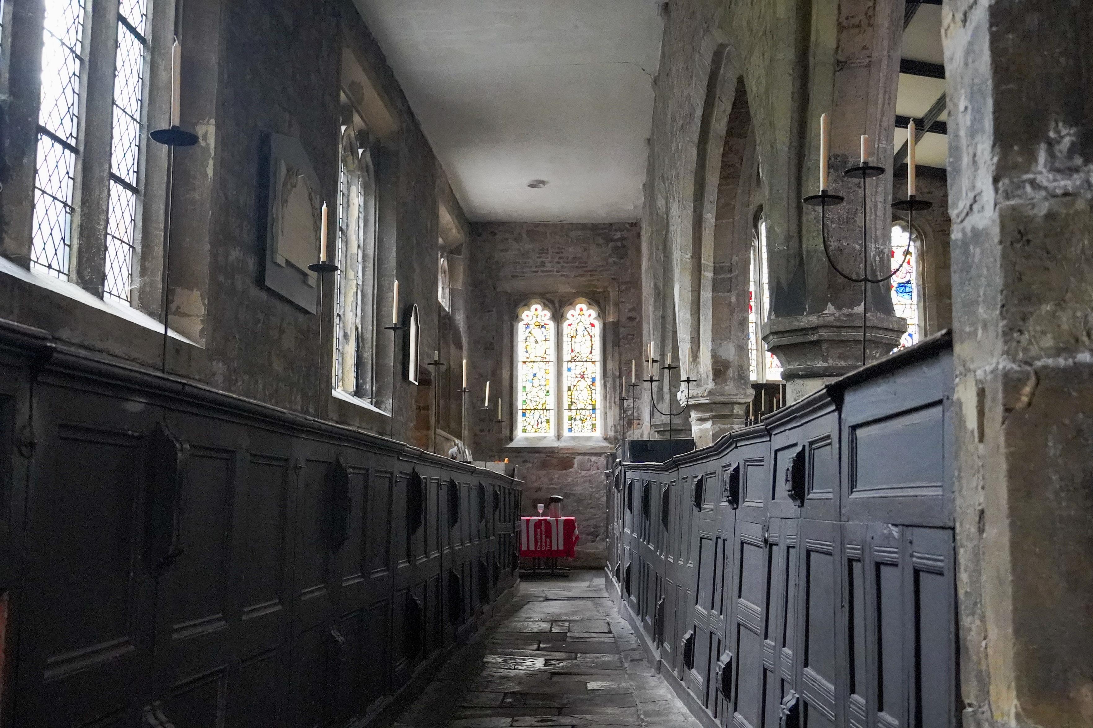The interior of an old stone church or chapel, viewed down a narrow flagstone aisle between two rows of dark wooden box pews. Tall leaded and stained-glass windows line the walls, with wrought-iron candle holders holding white candles. At the far end, a bright stained-glass window sits above a small table draped in a red cloth. The stonework is weathered and the light is soft and dim.