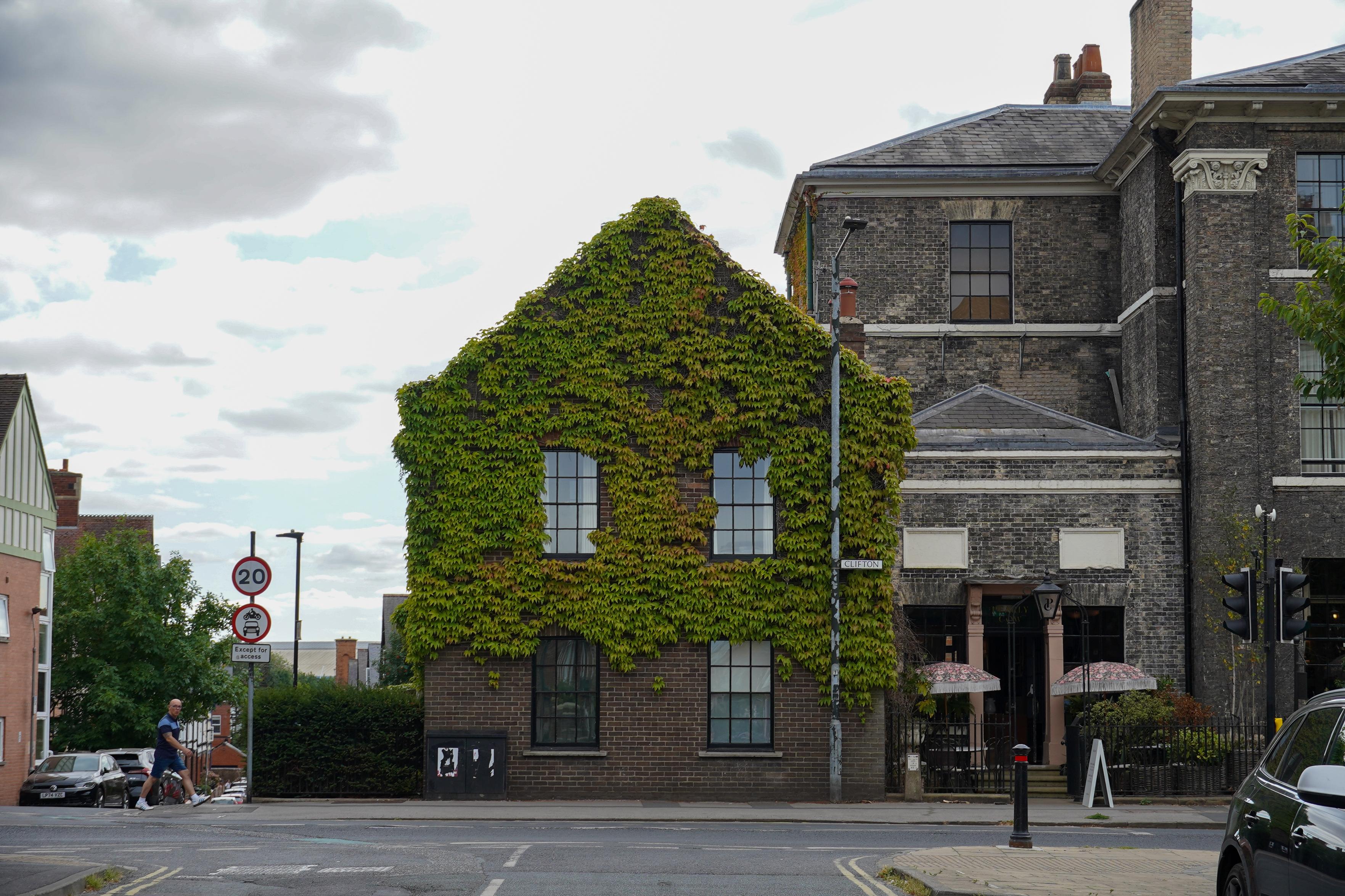 A street corner with a striking building whose entire gable end is covered in dense green ivy, almost swallowing the windows. Beside it stands a larger dark-brick Georgian building with tall sash windows and a pillared entrance shaded by fringed parasols. A street sign reads "Clifton", a 20 mph sign stands nearby, and a person in shorts walks along the pavement under a cloudy sky.