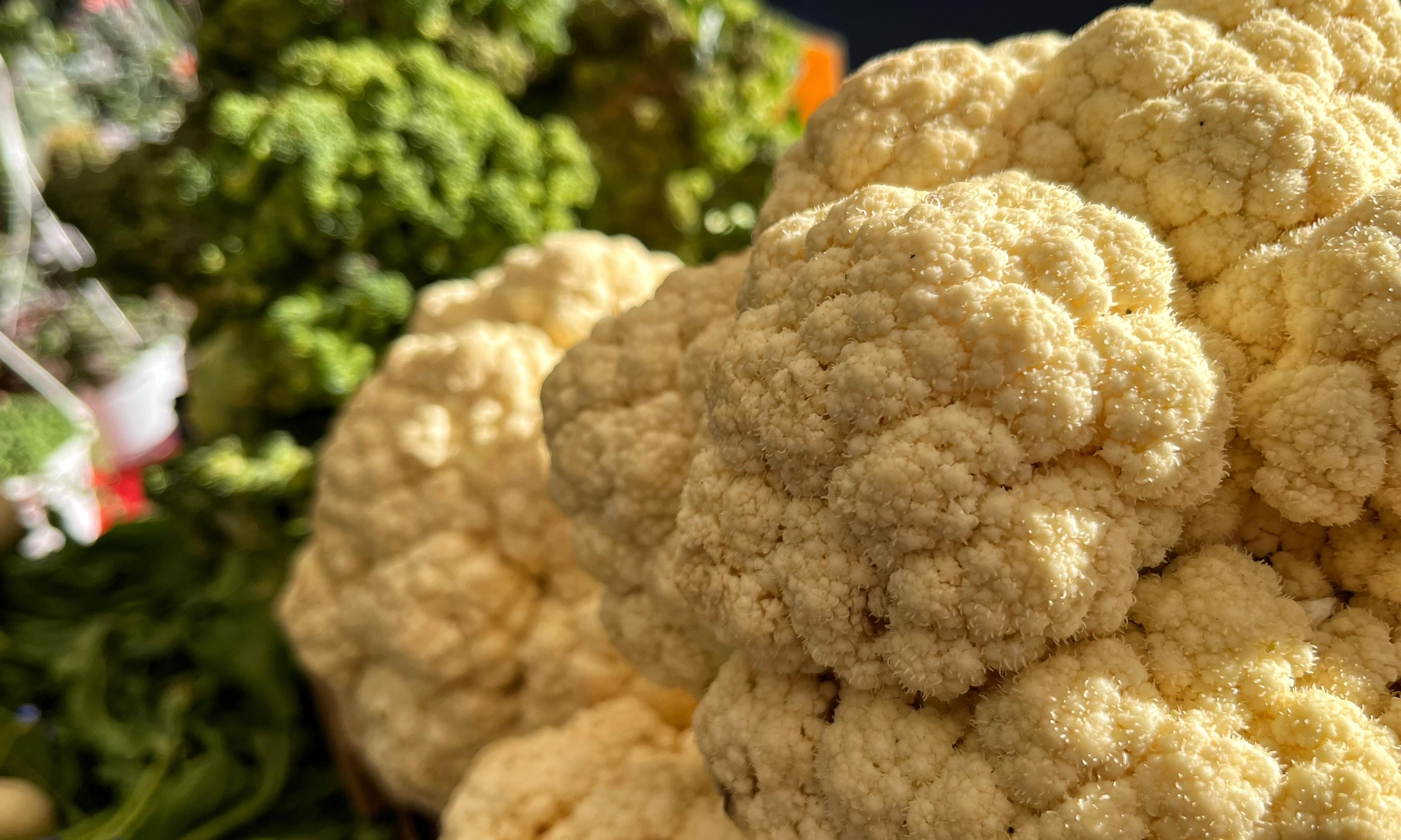 A large head of cauliflower with dense, white florets, side-lit by the morning sunshine at local farmers market, with green broccoli blurred in the background.