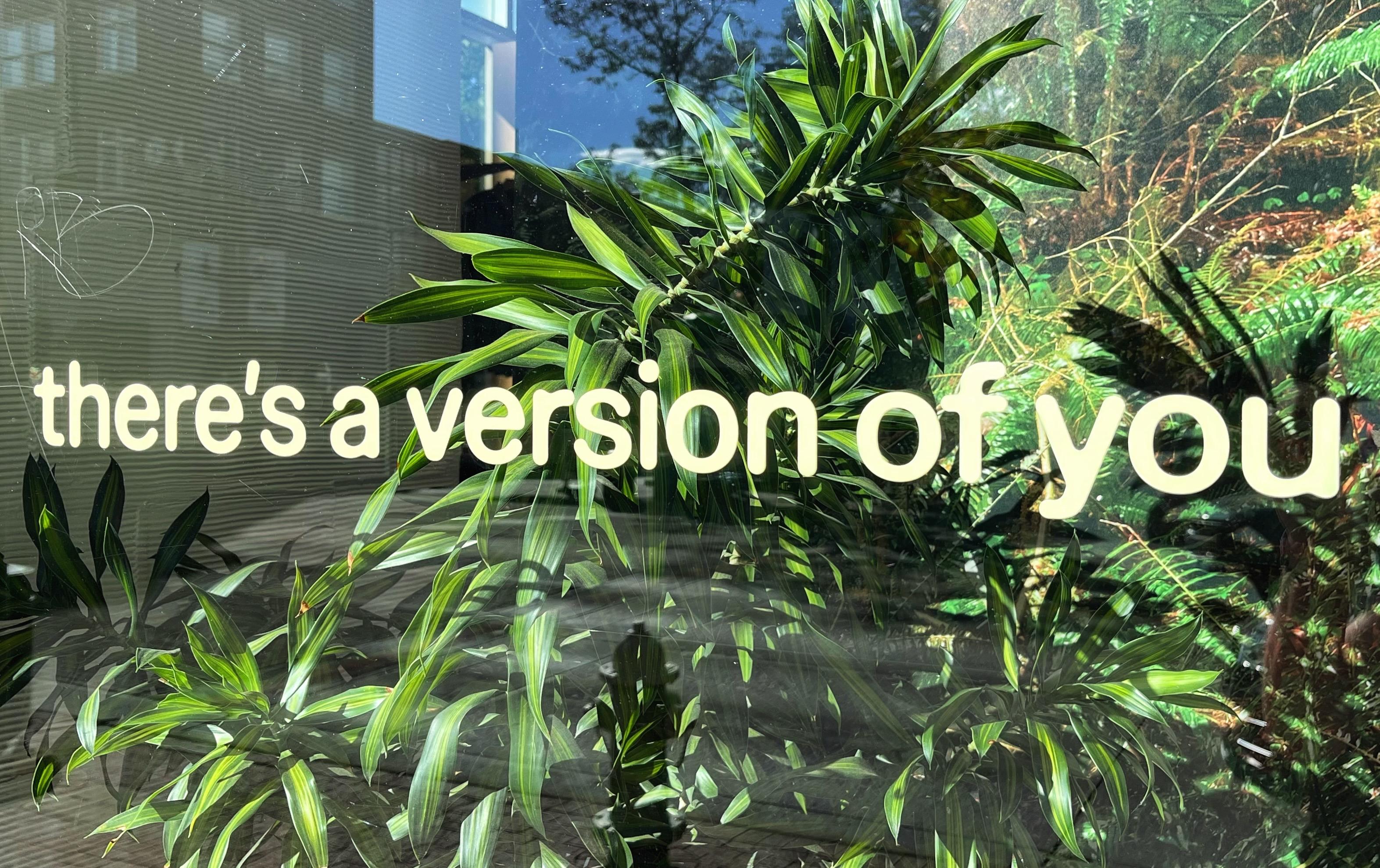 A large, glass display window in a downtown shop with green plants in the display and ghostly reflections of surrounding buildings on the glass. There are off-white, lowercase letters stuck to the glass that just say, “there’s a version of you.” It looks odd/strange/silly all by itself if you don’t notice that the rest of the sentence is pasted on the next two display windows.
