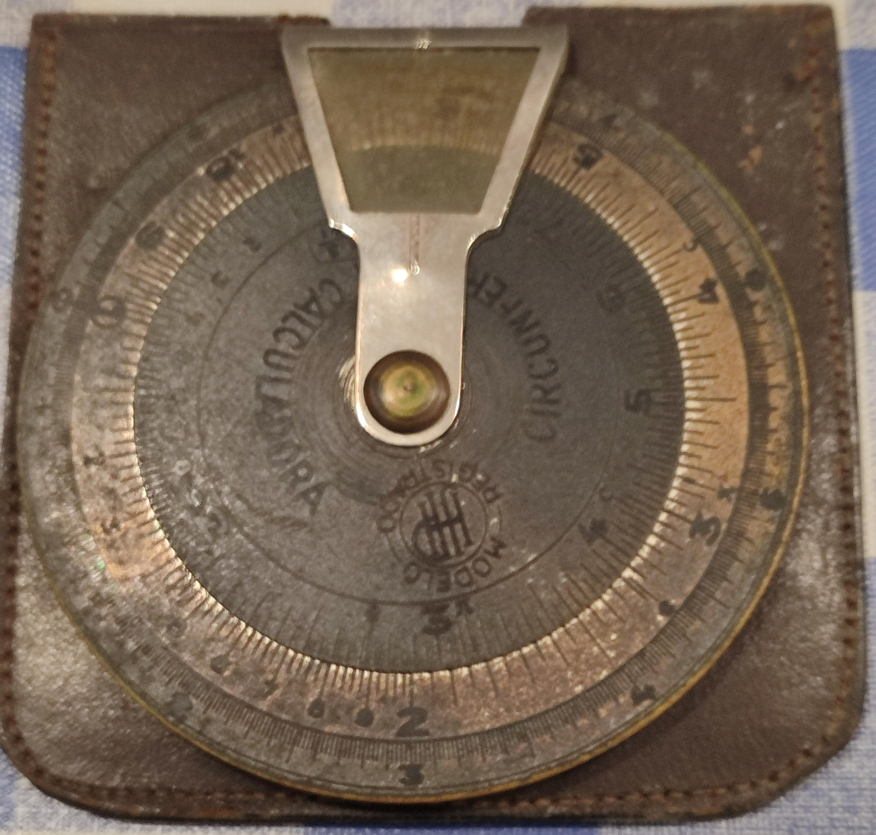 Circunferencia calculadora. Está compuesto por tres círculos concéntricos de diferentes aleaciones, más un marcador de metal y cristal para consultar el resultado del cálculo. Está puesta sobre su funda de cuero.